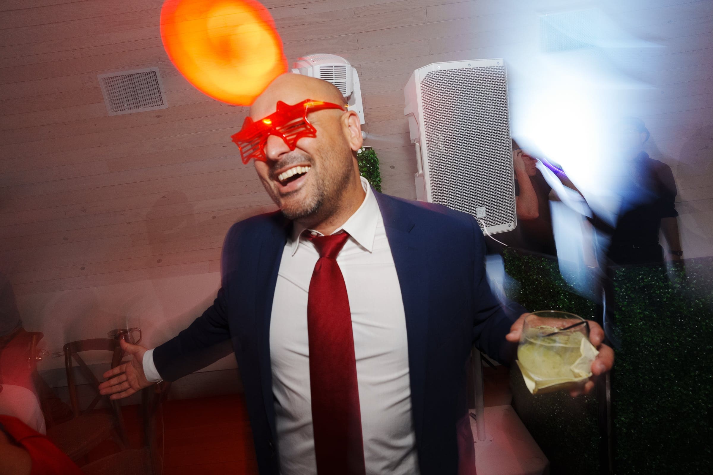 historic wakefield barn wedding - logan & rhett At a lively Wakefield Barn wedding, a man in a suit and red tie, wearing star-shaped red glasses, smiles widely. He holds a drink in one hand and seems to be enjoying himself. Blurred lights and a speaker enhance the festive atmosphere in the background.