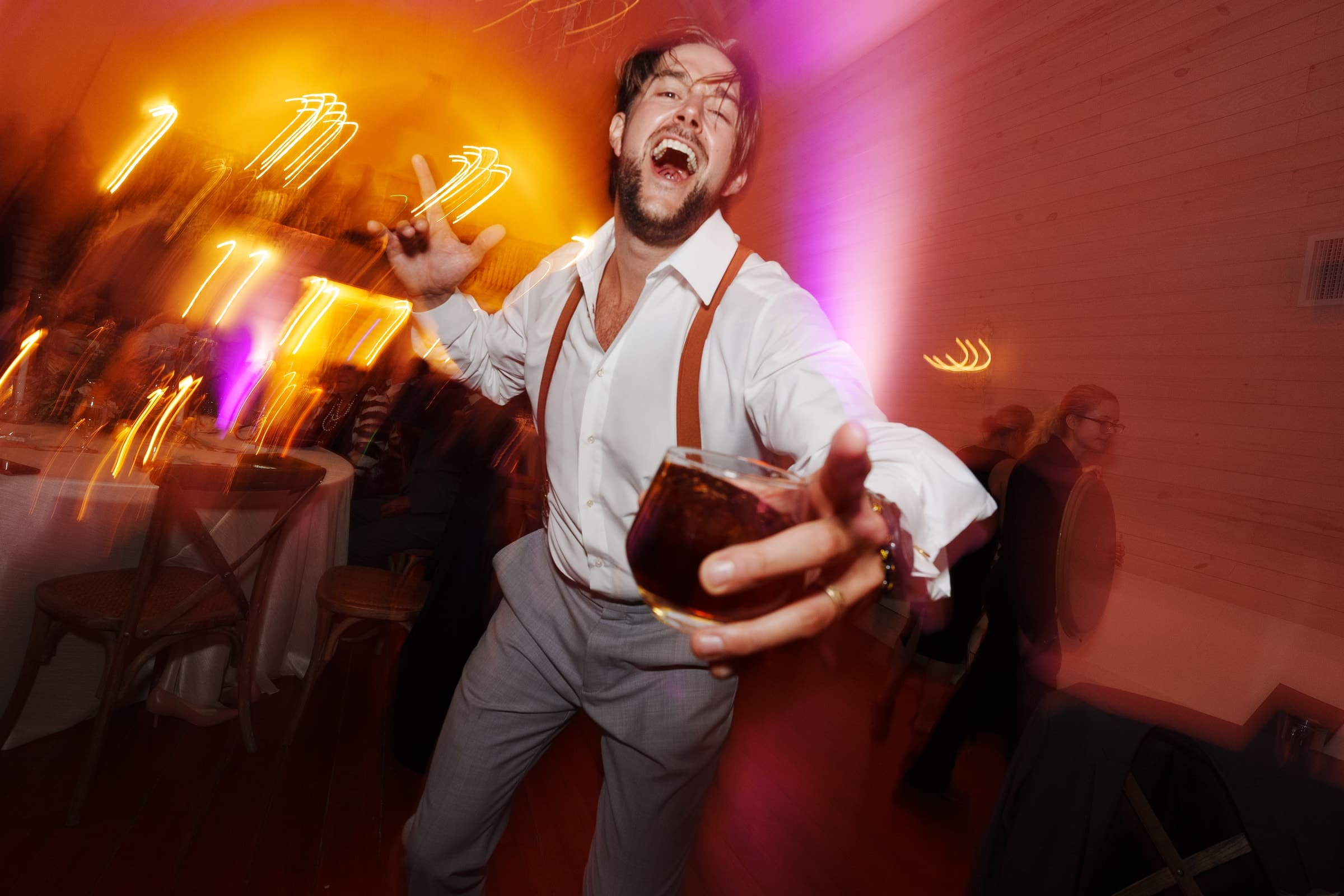 historic wakefield barn wedding - logan & rhett At a lively indoor party at Wakefield Barn, a man in a white shirt and suspenders dances joyfully. With a drink in one hand and flashing a peace sign with the other, he's surrounded by warm, glowing lights and blurred motion, perfectly capturing the festive wedding atmosphere.