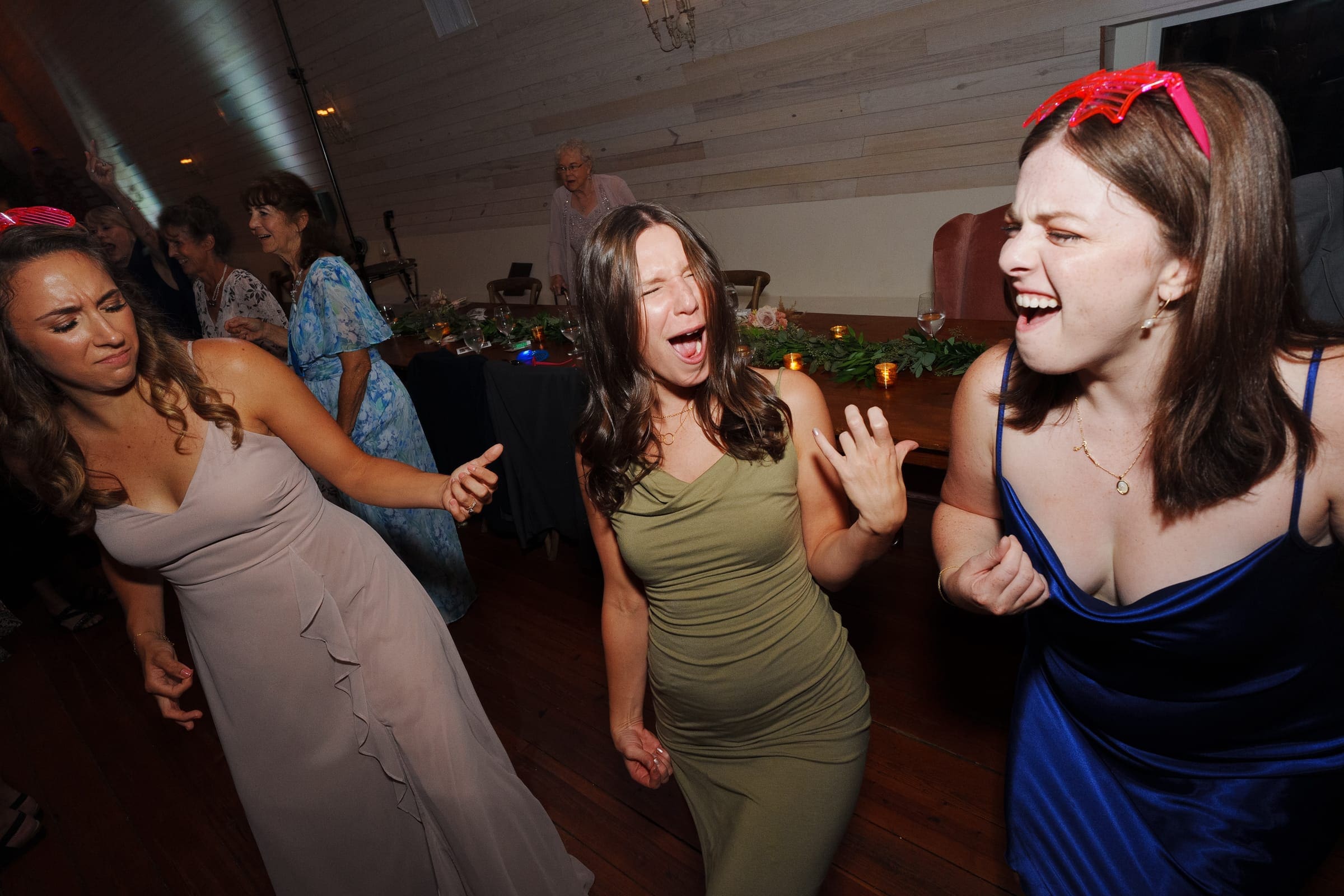 historic wakefield barn wedding - logan & rhett Three women are dancing energetically at a lively indoor party, clad in colorful dresses, reminiscent of a Wakefield Barn wedding. The background features wooden paneling and blurred guests, creating an atmosphere that's both joyful and festive.