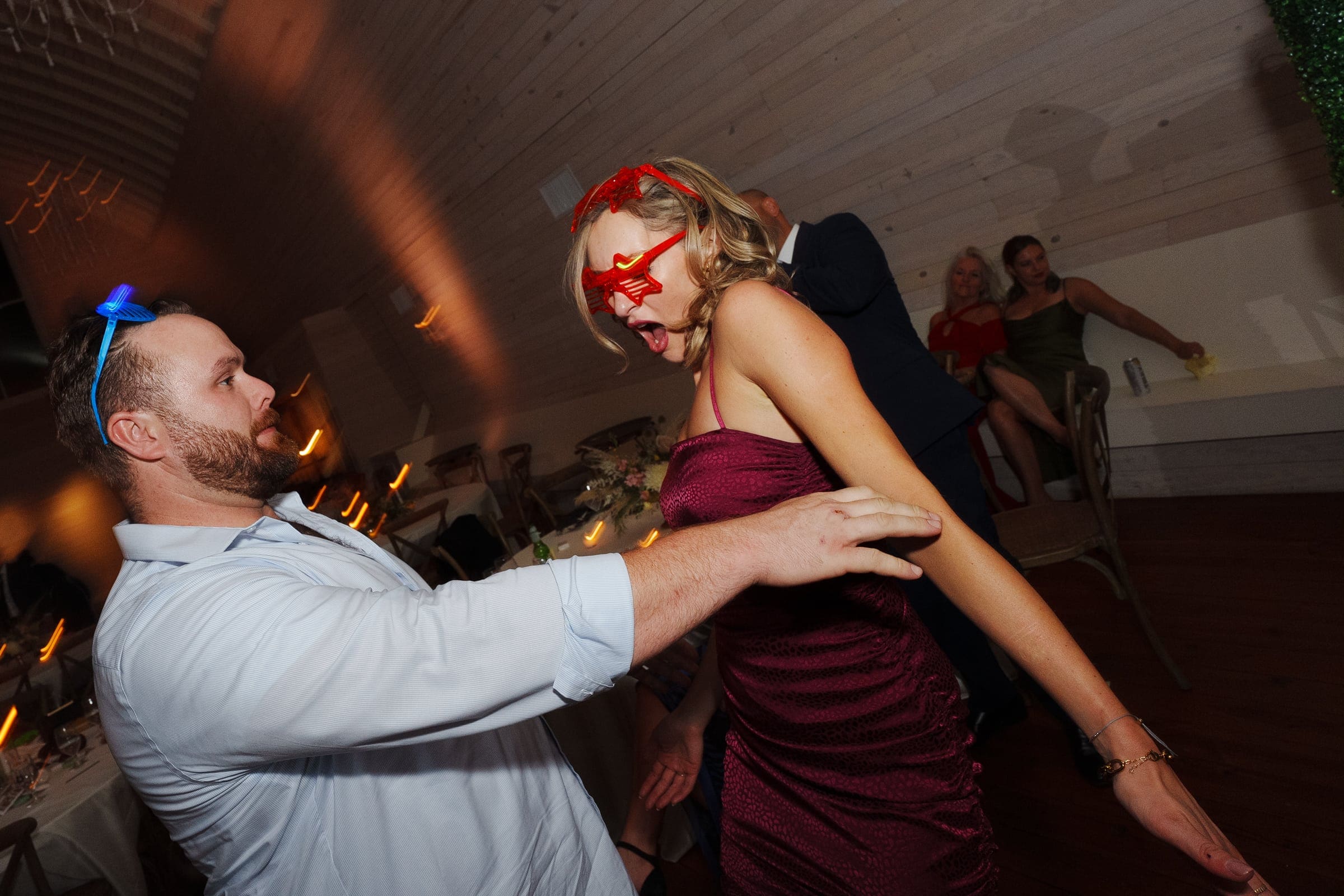 historic wakefield barn wedding - logan & rhett A man and woman are dancing energetically at a Wakefield Barn wedding, with the man wearing a blue bow headband and the woman in a red dress and mask. They seem to be having fun, with blurred movement around them and dim lighting enhancing the lively atmosphere.