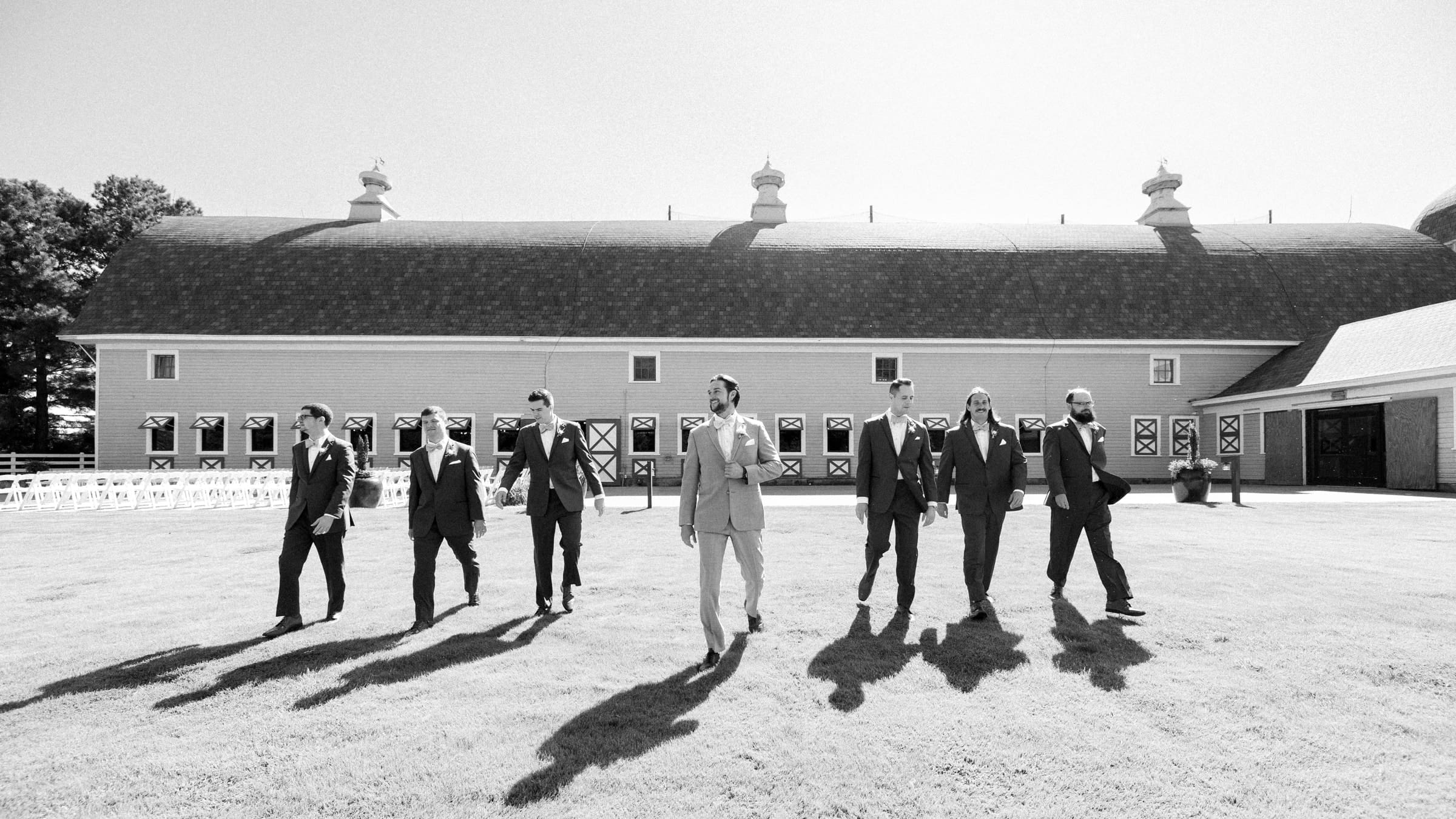 historic wakefield barn wedding - logan & rhett A group of eight people in suits strolls across a grassy field at the Wakefield Barn wedding, their long shadows stretching under the sun. The barn's classic architecture sets a formal and festive atmosphere.