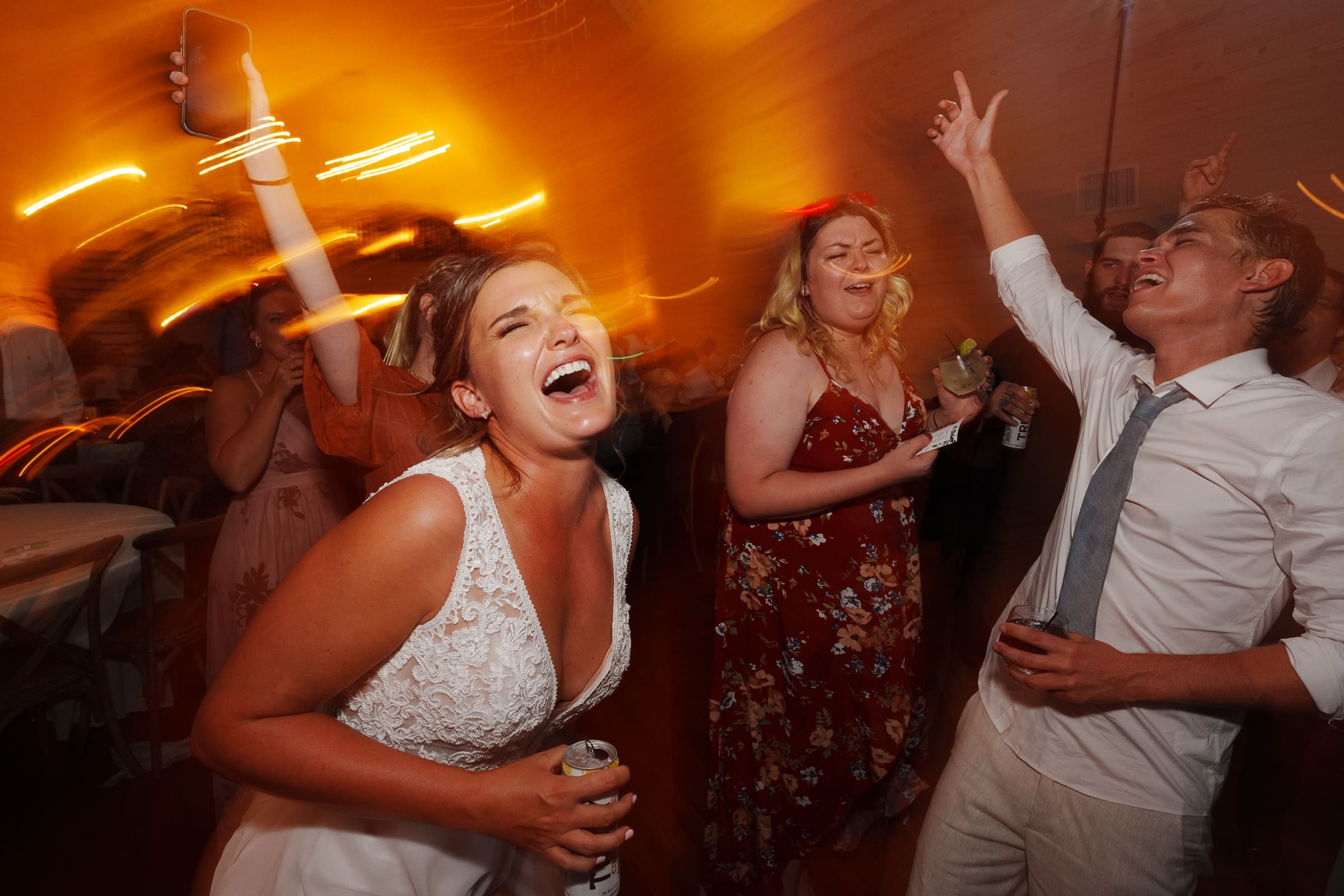 historic wakefield barn wedding - logan & rhett A joyful bride dances energetically at a lively Wakefield Barn wedding, surrounded by friends. The room glows with warm, orange lighting. People have drinks in hand and expressions of excitement, capturing the festive atmosphere.