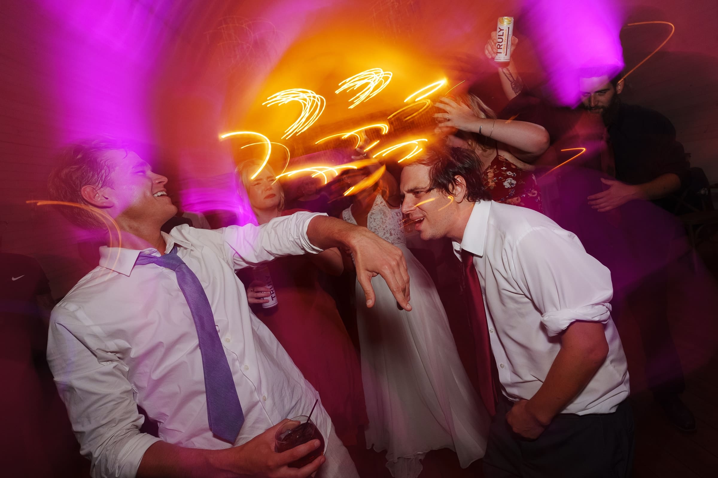 historic wakefield barn wedding - logan & rhett A lively scene captures a group of people dancing under vibrant purple and orange lights at the Wakefield Barn wedding. Two men in the foreground enjoy the music, one holding a drink. In the background, others with raised arms and dynamic motion blur create an energetic atmosphere.