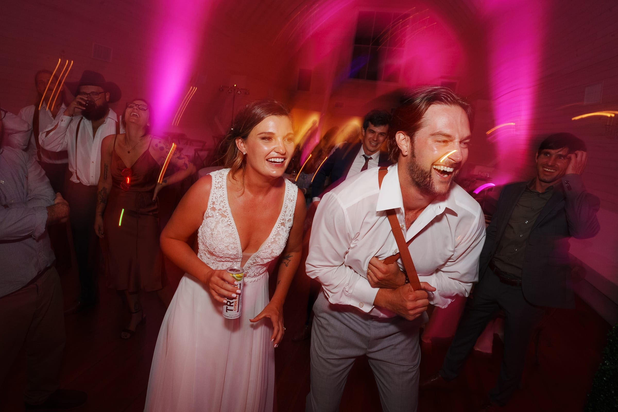 historic wakefield barn wedding - logan & rhett At a Wakefield Barn wedding, a bride and groom laugh joyfully on a vibrant dance floor, surrounded by friends. Colorful party lights illuminate the scene, enhancing the lively atmosphere. The groom holds a guitar strap while the bride carries a drink can, both in their elegant wedding attire.