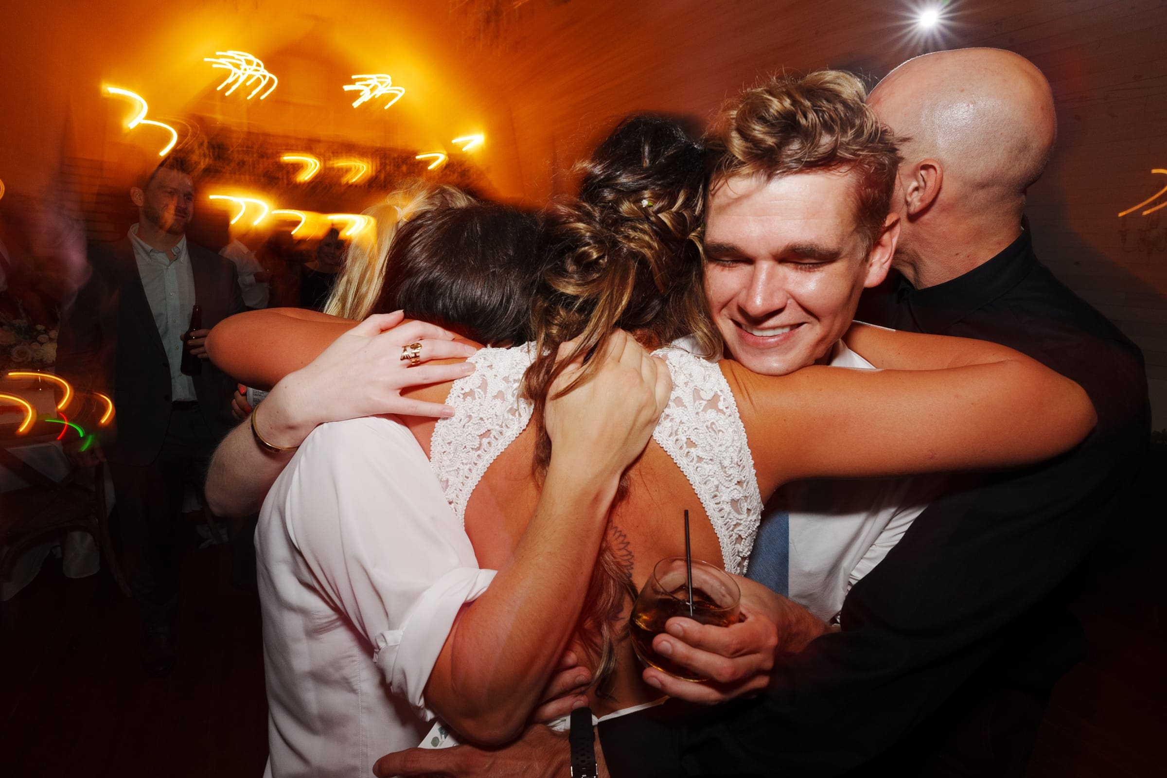 historic wakefield barn wedding - logan & rhett At a lively Wakefield Barn wedding, a group of people huddles together in a warm embrace. The atmosphere is filled with motion and joy, with blurred lights creating an energetic background. One person holds a drink, and everyone seems to be celebrating the special occasion.