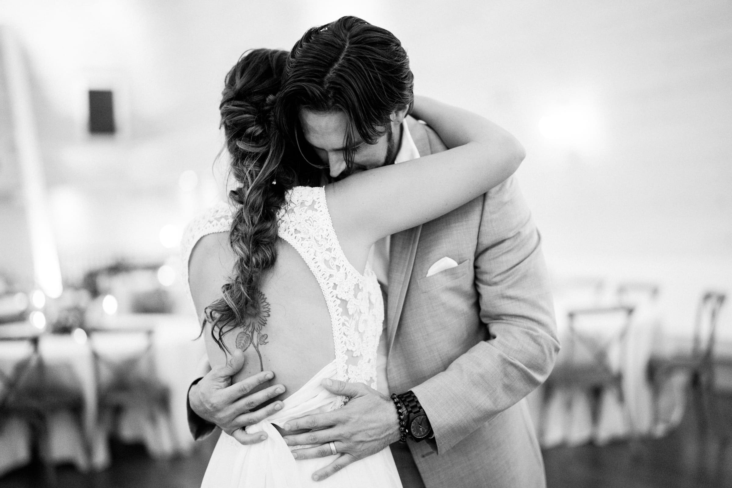 historic wakefield barn wedding - logan & rhett At a Wakefield Barn wedding, a couple embraces on the dance floor. The woman, her long hair in a braid, wears a sleeveless dress with lace detailing while the man dons a light-colored suit. A softly blurred background enhances the intimate atmosphere.