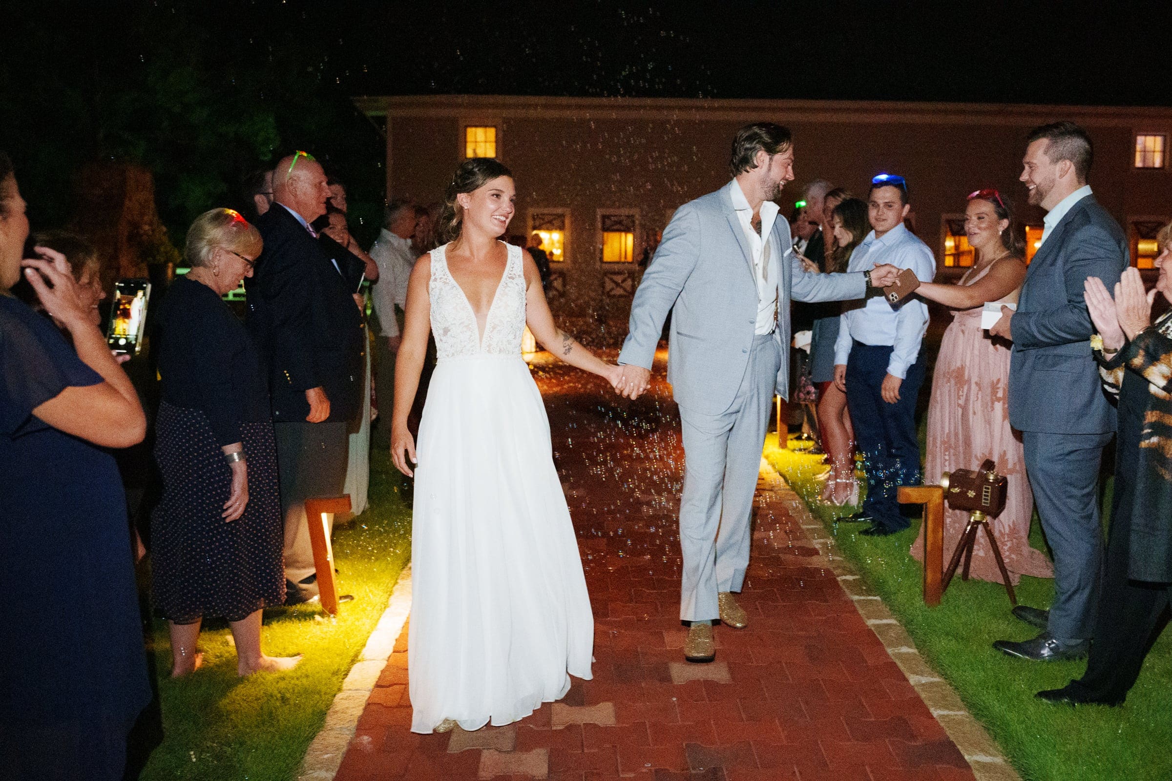 historic wakefield barn wedding - logan & rhett A bride and groom hold hands, walking down the path at their Wakefield Barn wedding, surrounded by smiling guests in formal attire who clap and throw confetti into the night. The bride dazzles in a white dress, while the groom looks sharp in a light gray suit.
