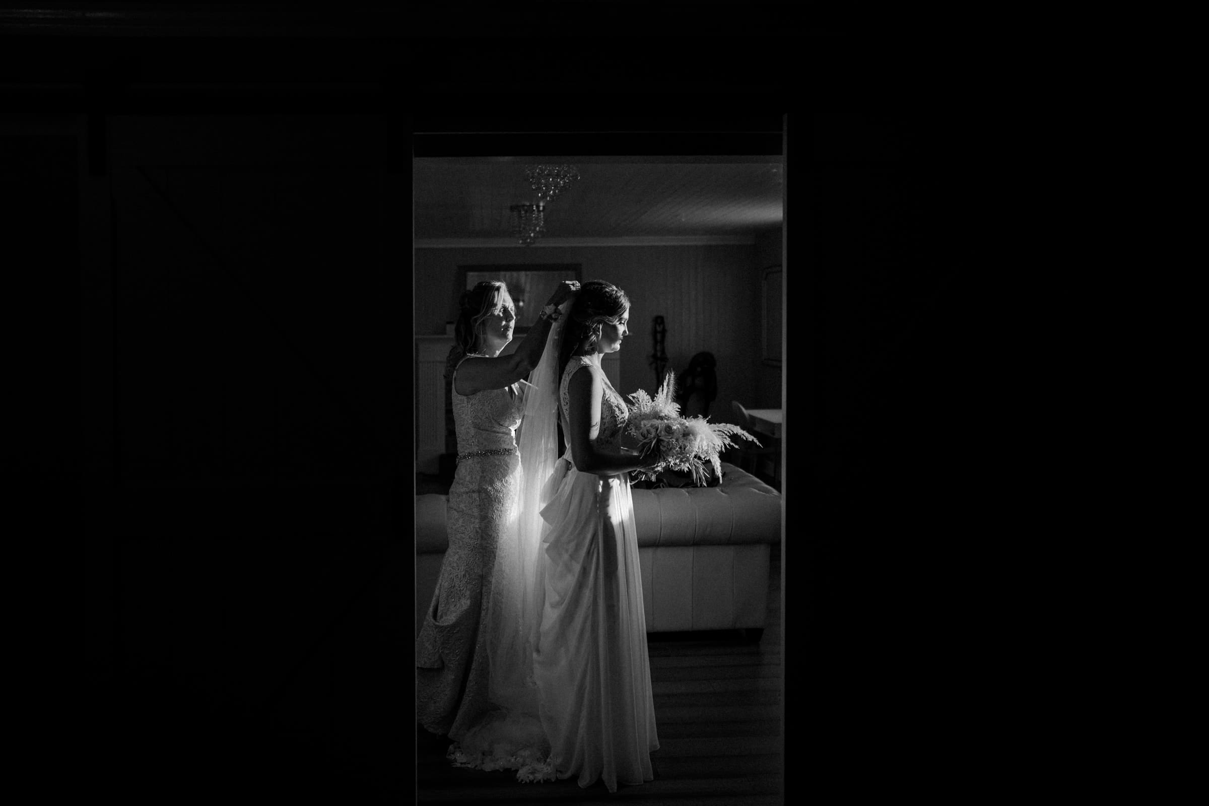 historic wakefield barn wedding - logan & rhett A bride stands in profile with a bouquet at a Wakefield Barn wedding, while another woman adjusts her hair. They are in a dimly lit room, framed by the doorway, with soft light highlighting their faces and dresses.