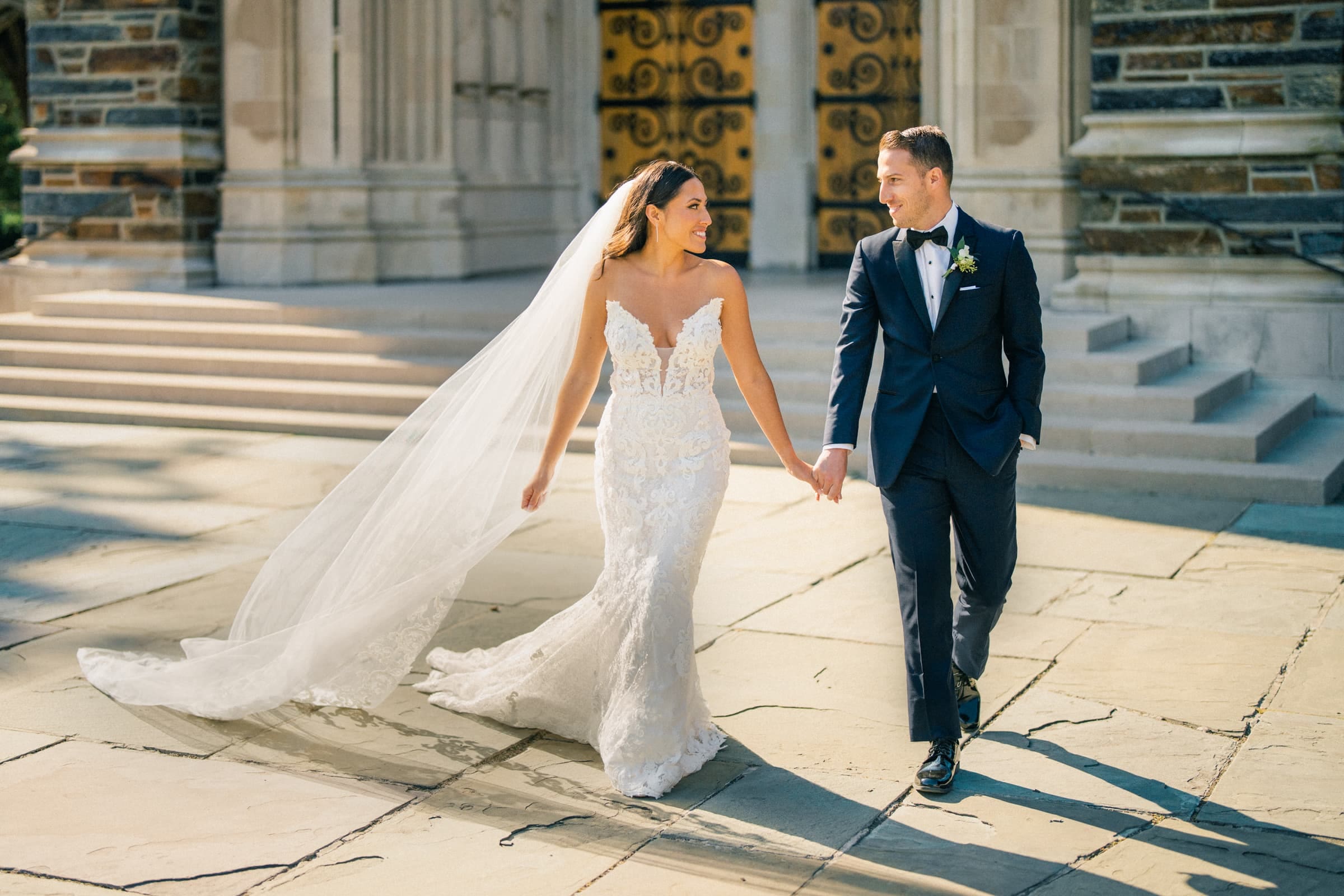 A joyful wedding portrait of a bride and groom smiling at each other while walking hand-in-hand on a sunny path outside Duke Chapel, featuring the grand building and ornate doors.