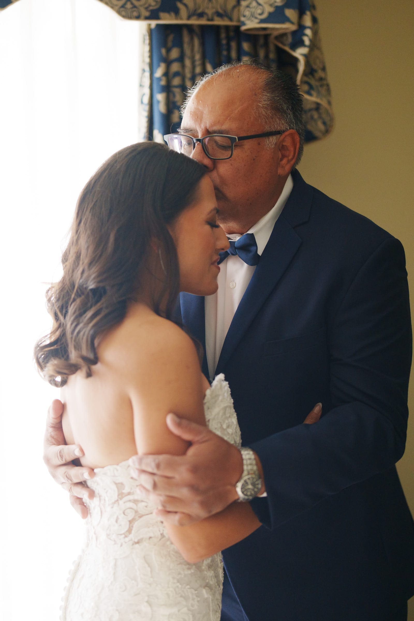 A man in a suit gently embraces a woman in a white dress, kissing her forehead amid the softly lit ambiance of Duke Gardens. Patterned curtains frame the background, enhancing the scene's sense of tenderness and affection on their special wedding day.