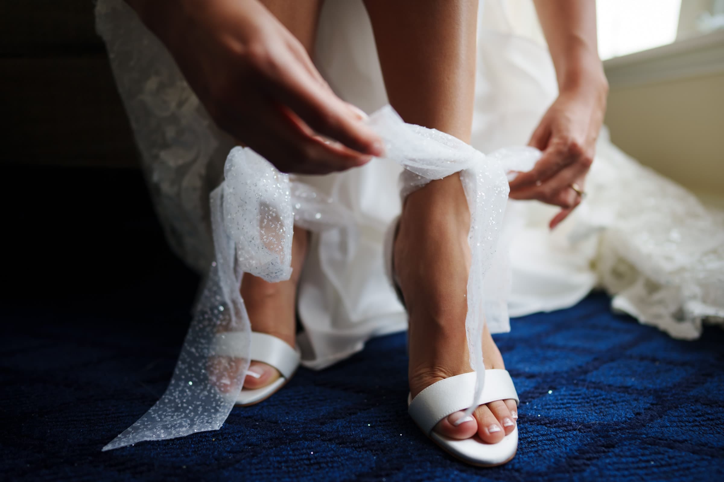 A person in a white dress is tying sheer, glittery ribbons around their ankles over white heels, as if preparing for a Duke Gardens wedding, while sitting on a blue carpet.