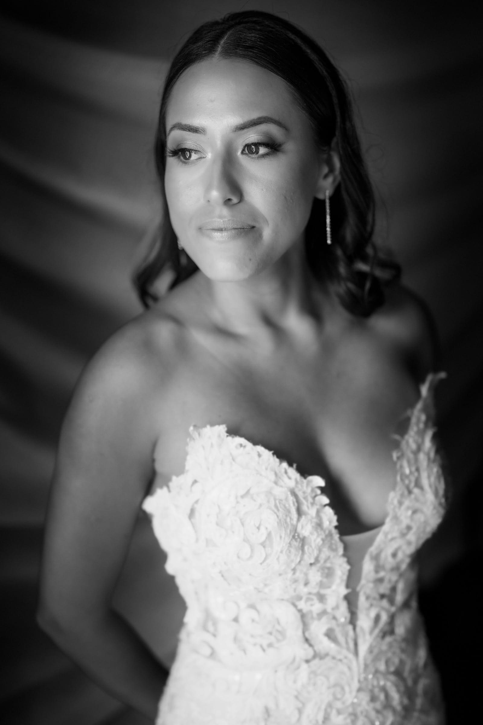Black and white portrait of a bride looking pensive in Duke Gardens. She is wearing an elaborate lace wedding dress with thin straps and drop earrings. The background is softly blurred, emphasizing her serene expression.