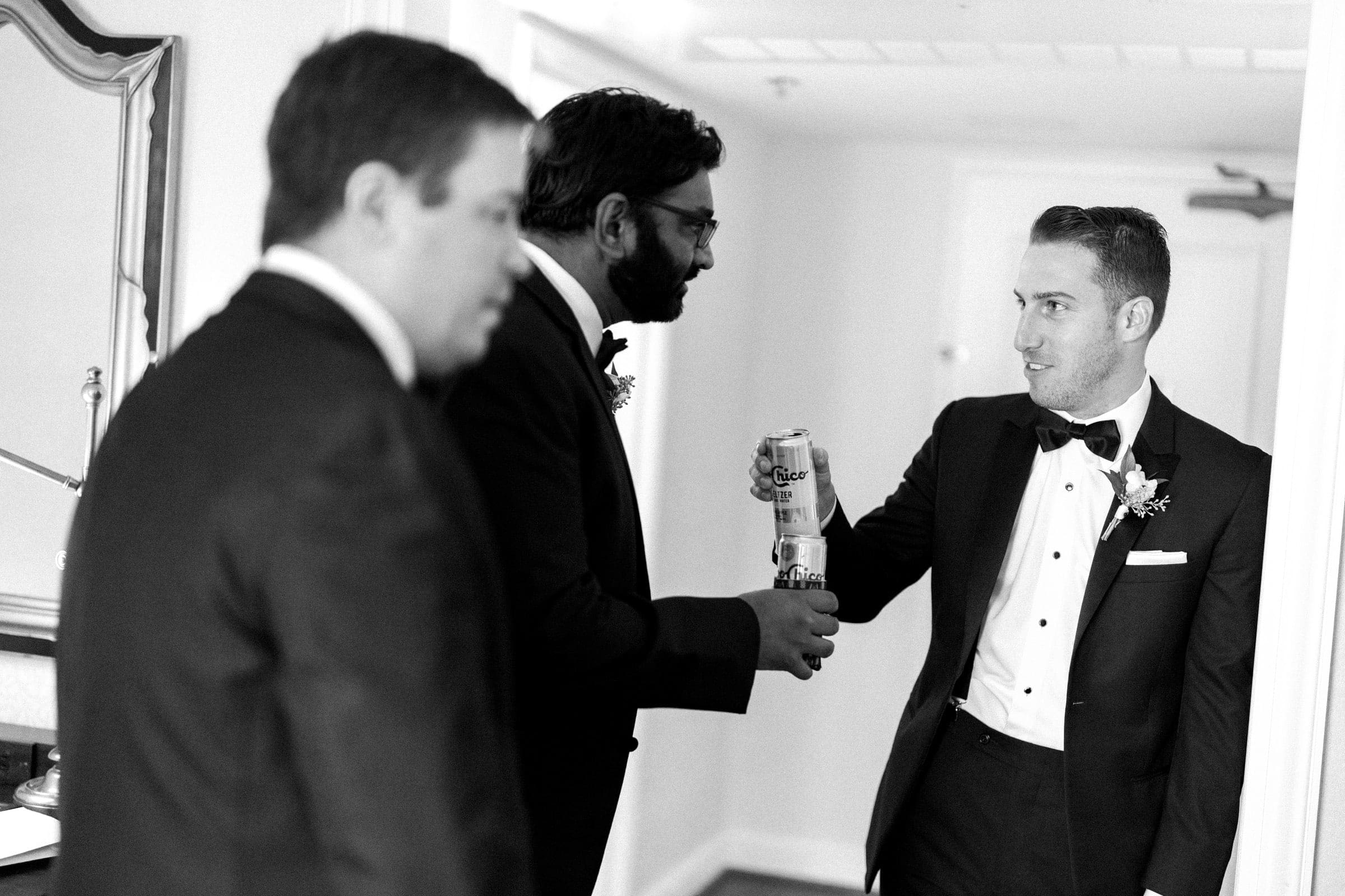 Three men in suits stand in a room, two holding beer cans as they toast with smiles. They appear to be preparing for a Duke Gardens wedding. The scene exudes a relaxed and celebratory mood, perfectly setting the tone for the elegant event ahead.