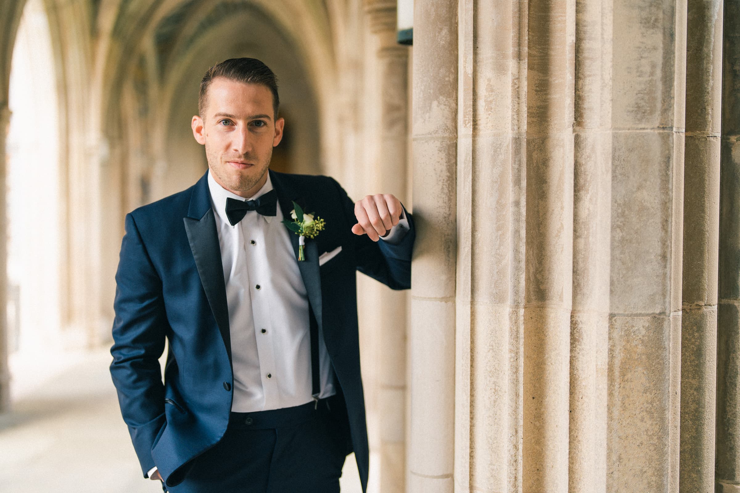 A man in a navy suit with a bow tie stands confidently, one hand in his pocket and the other resting on a stone column, as if ready for a Duke Gardens wedding. A boutonniere is pinned to his lapel, perfectly complementing the elegant hallway with its arched ceilings.