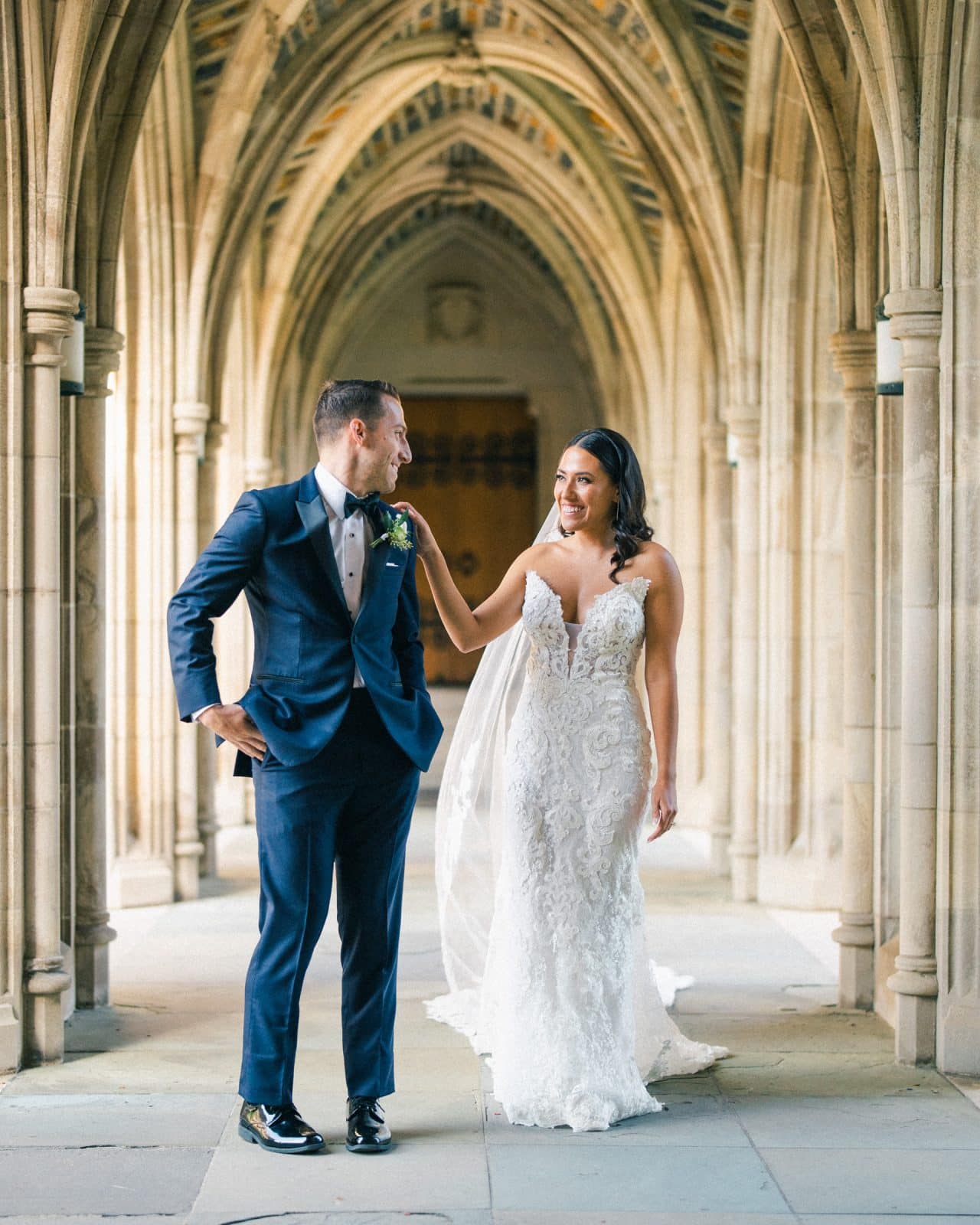 A candid fine art wedding photograph capturing a first look in the stone arcades of Duke Chapel, showing a Latina bride placing her hand on the groom's shoulder as he turns to see her.