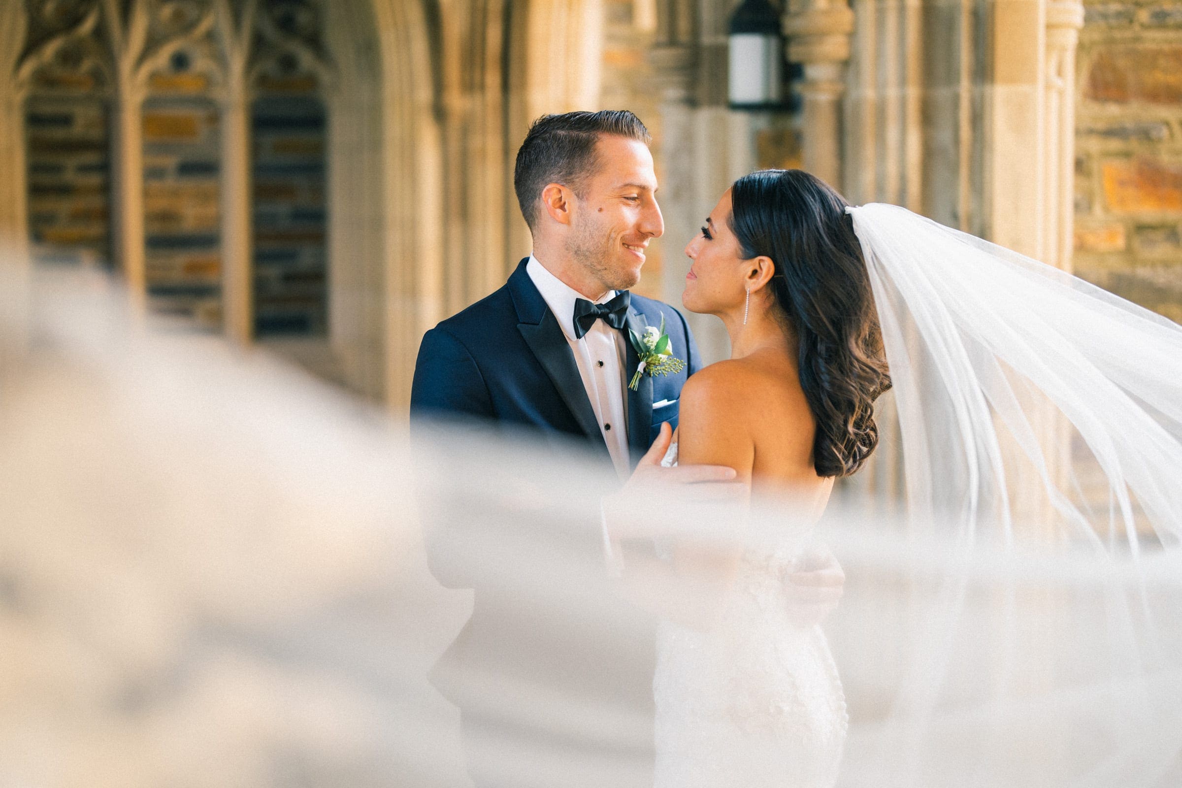 A bride and groom share a romantic moment in the elegant setting of Duke Gardens. The bride's long veil flows in the foreground as they stand dressed in formal wedding attire, gazing into each other's eyes with warm smiles.