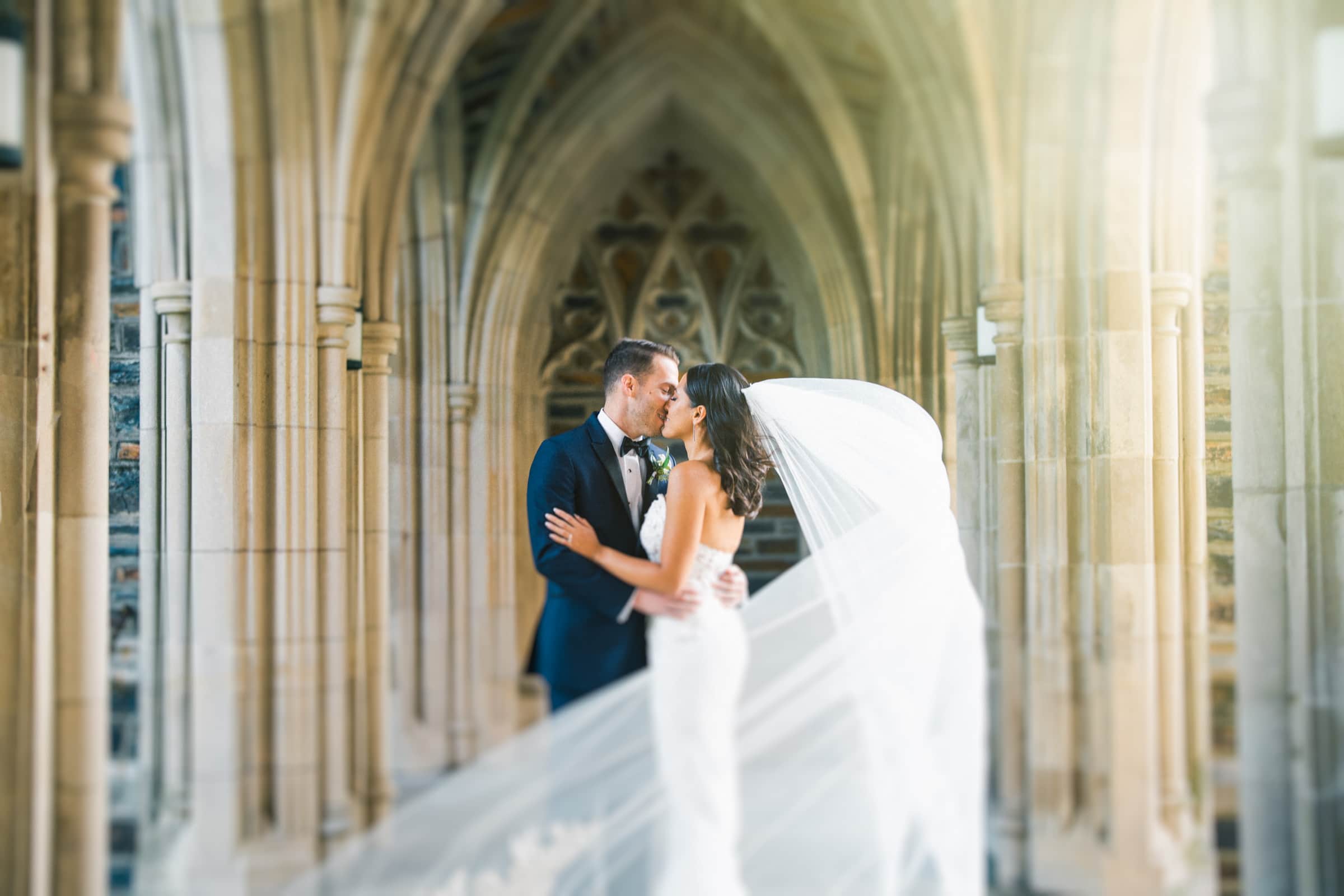 A bride and groom share a kiss under a stone archway at Duke Gardens. The bride wears a flowing dress with a long veil, and the groom is in a dark suit. Sunlight filters in, creating a romantic and serene atmosphere for their wedding.