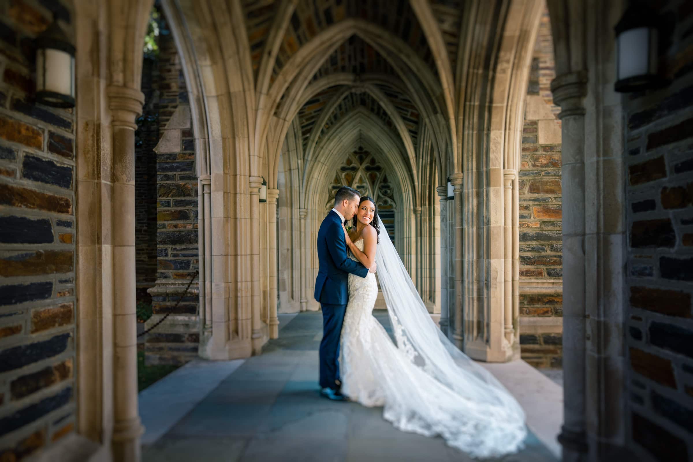A bride and groom stand closely under a stone archway at Duke Gardens, embracing. The bride wears a long, flowing veil and a lace gown, while the groom is in a dark suit. Sunlight softly illuminates the architectural details and the couple's attire in this enchanting wedding scene.