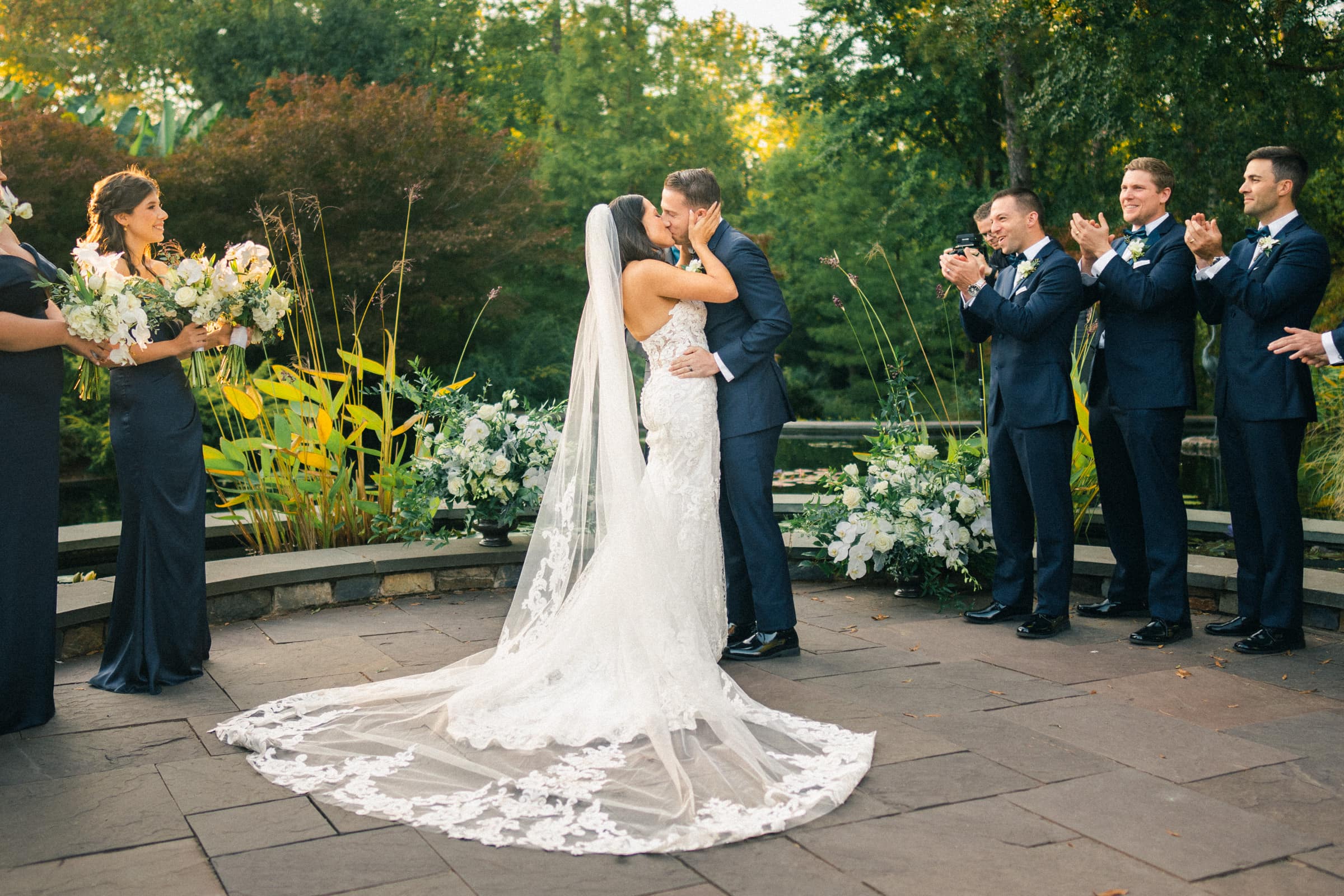 A bride and groom kiss during their Duke Gardens wedding ceremony outdoors. The bride wears a long, lace gown with a veil, and the groom is in a dark suit. Bridesmaids and groomsmen stand by with bouquets, clapping amidst lush greenery and floral arrangements.