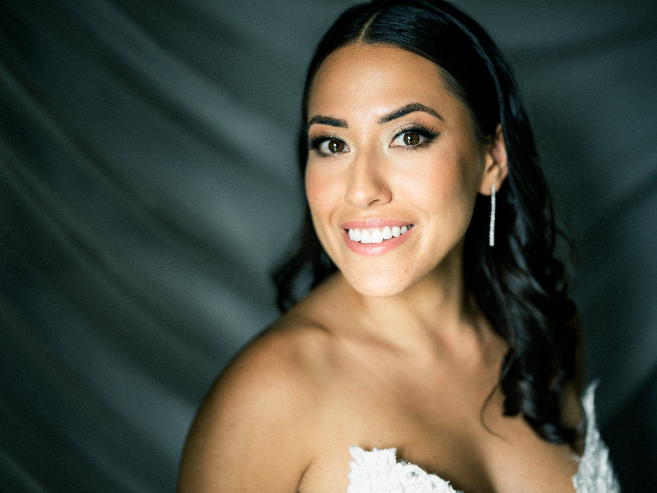 A woman smiling warmly at the camera, wearing a strapless white dress, exudes elegance reminiscent of a Duke Gardens wedding. Her long, dark hair is styled in loose curls and she wears dangling earrings. The background is a soft, blurred gray fabric.