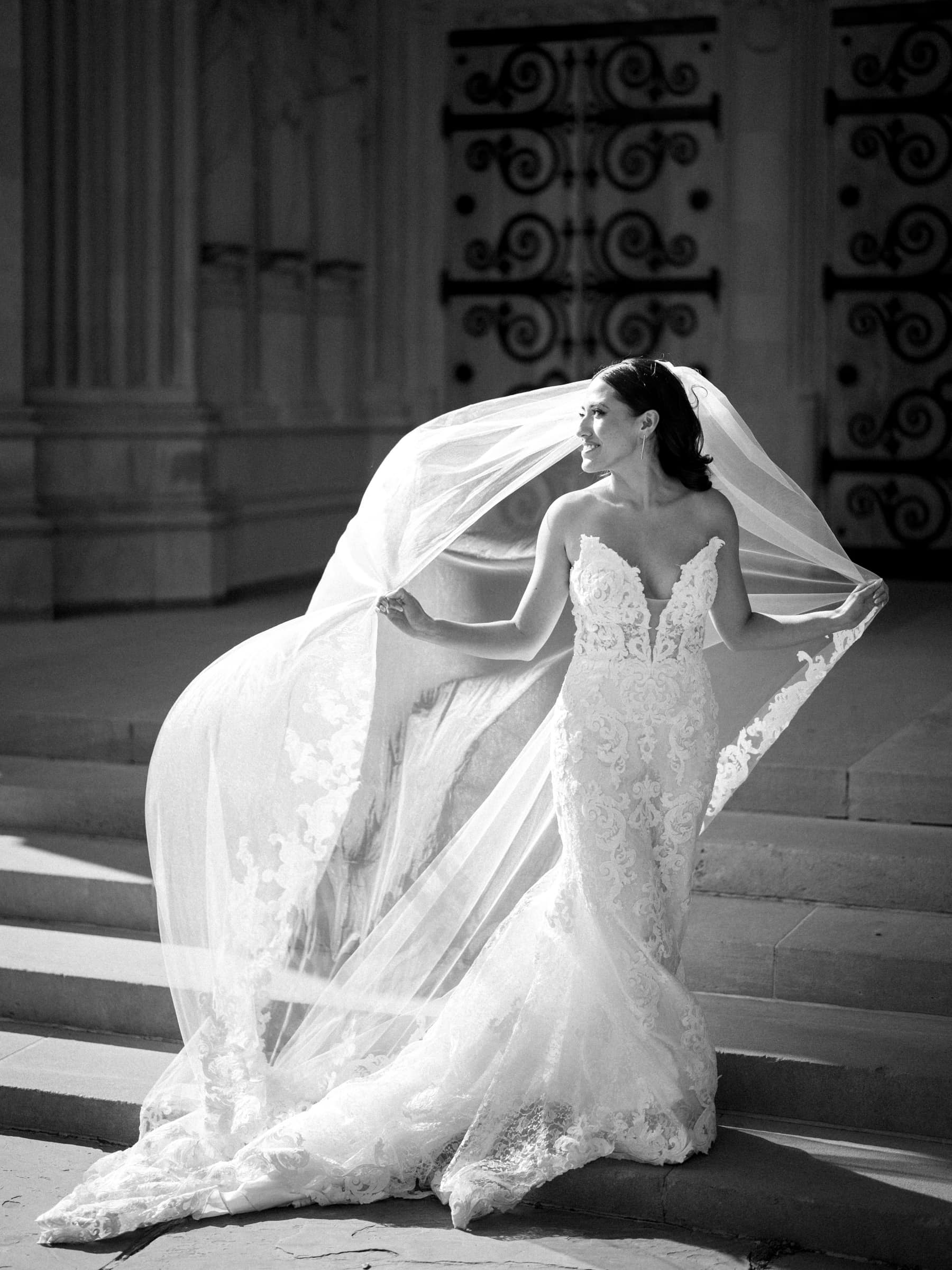 A bride in an intricate lace wedding dress stands on stone steps at Duke Gardens, holding her long veil as it flows in the sunlight. She gazes to the side, set against ornate doors in the background.