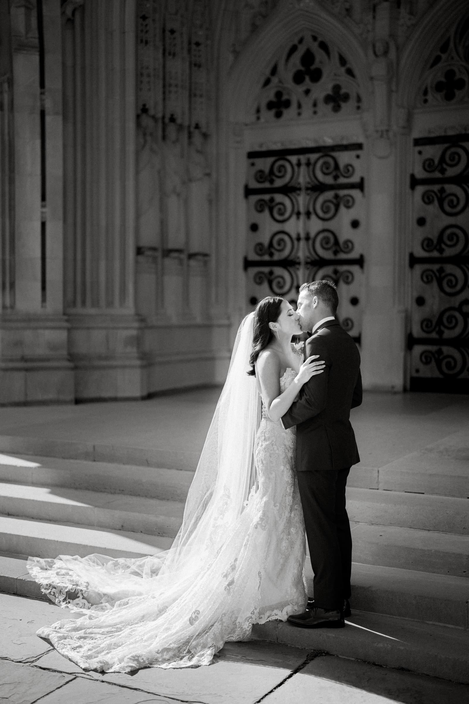 A black-and-white photo captures a couple embracing on the steps of a grand building, perhaps reminiscent of a Duke Gardens wedding. The bride, in a flowing gown with a long veil, stands beside the groom in his suit. Ornate doors with intricate patterns frame this timeless moment.