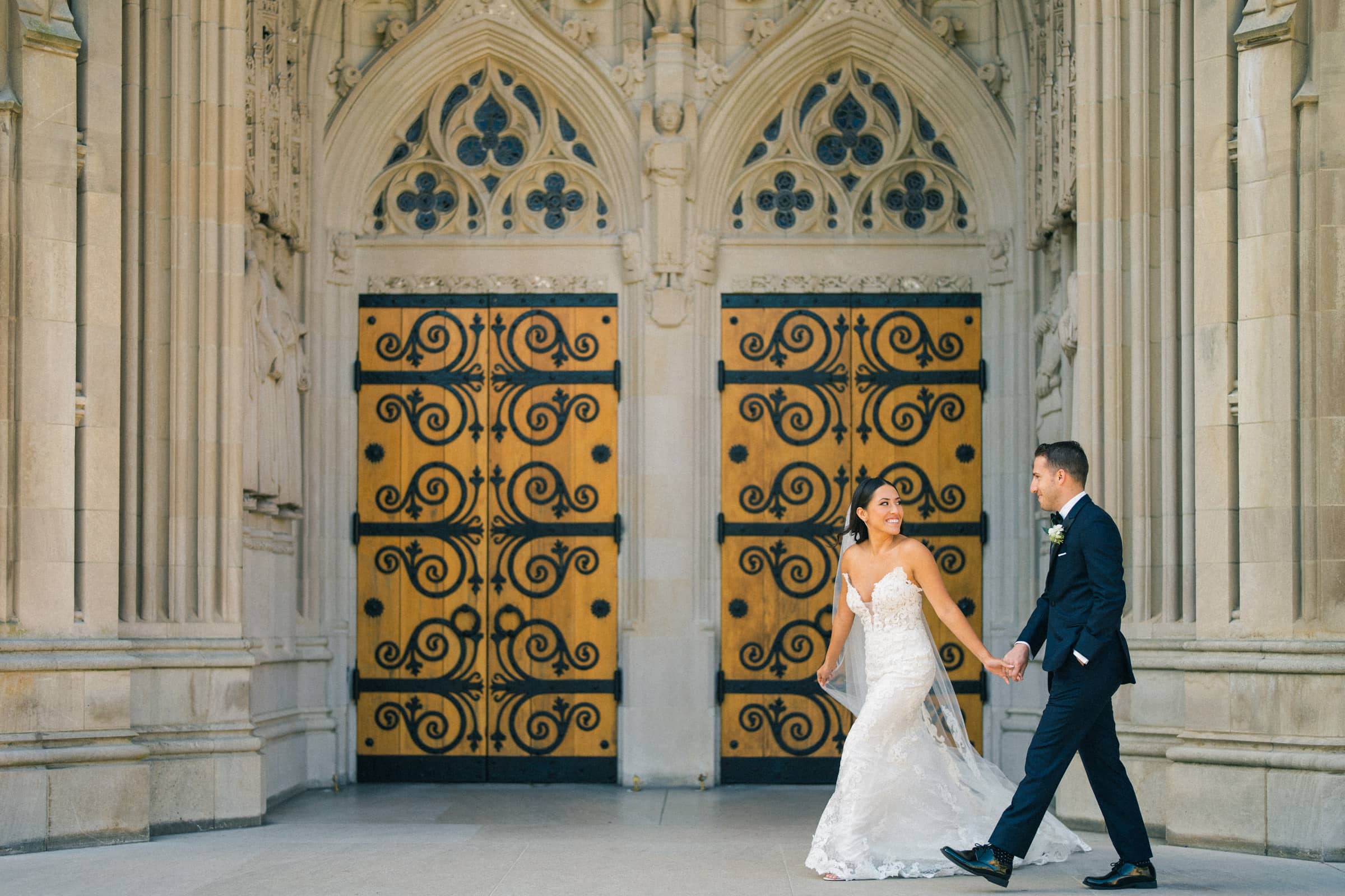 A bride and groom hold hands and smile at each other while walking in front of a grand, ornate building with large, decorative doors at Duke Gardens. The bride wears a lace gown, and the groom is in a dark suit.