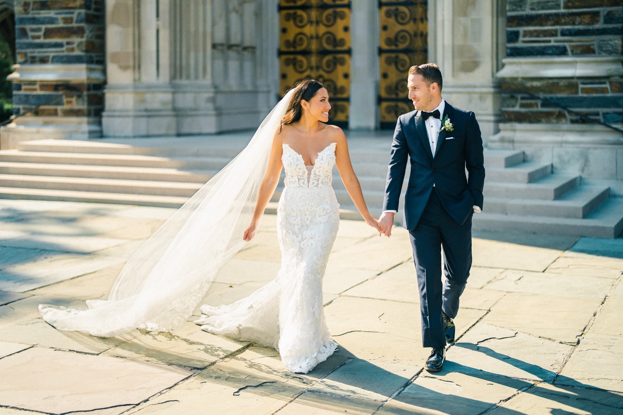 A bride and groom hold hands, walking outside a stone building at Duke Gardens. The bride wears a white lace gown with a long veil, and the groom is in a dark suit with a boutonniere. Both are smiling and looking at each other.