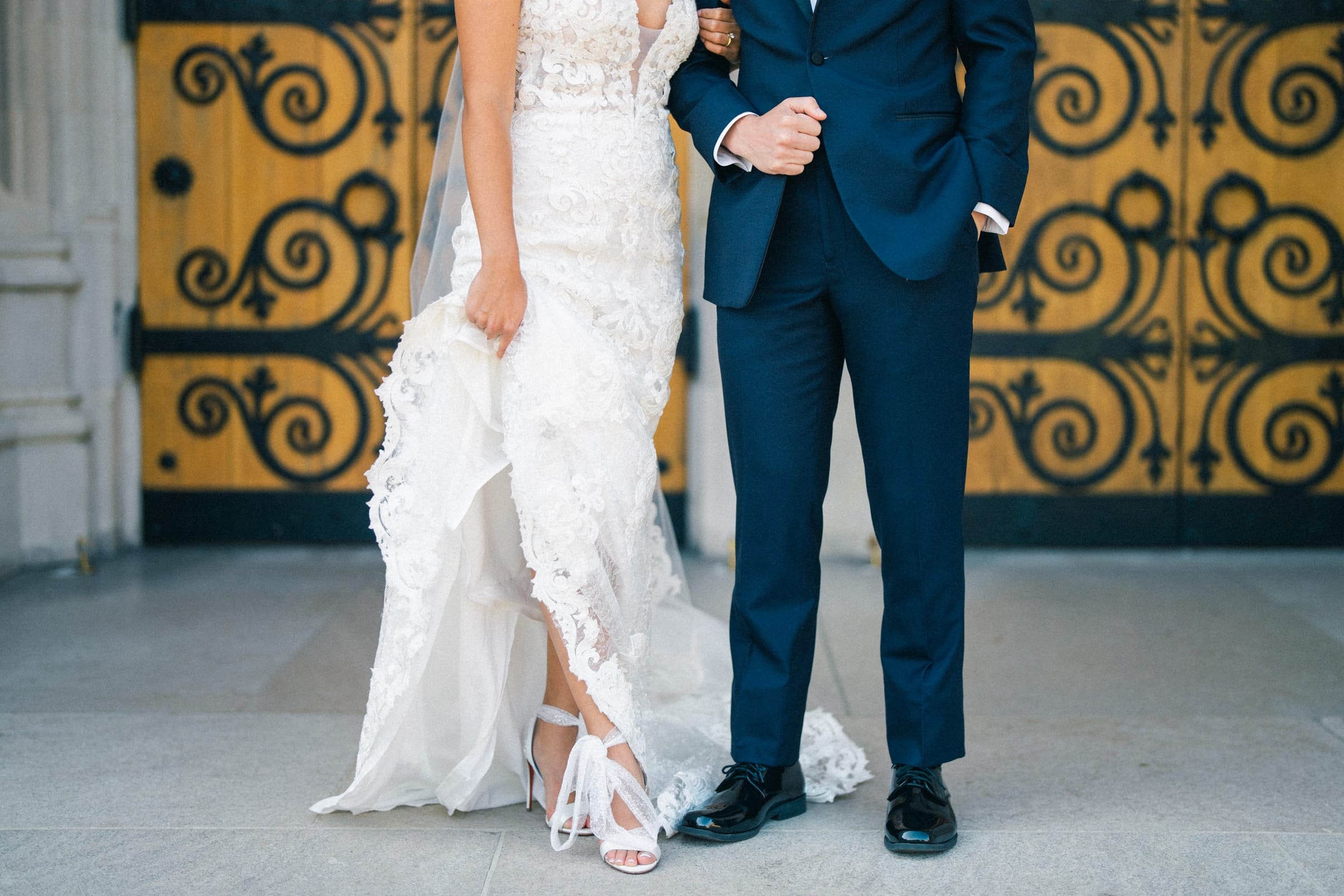 A bride in a white lace wedding dress and heels holds her skirt, standing arm in arm with a groom in a navy blue suit and black shoes. They grace the entrance of ornate wooden doors with black iron swirls, capturing the elegance of a Duke Gardens wedding.