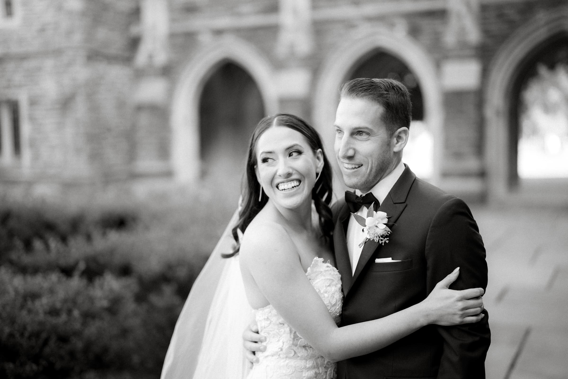A black-and-white photo captures a smiling bride and groom embracing outside a stone building with arched windows, reminiscent of a Duke Gardens wedding. The bride wears a lace dress and veil, while the groom dons a suit with a boutonniere.