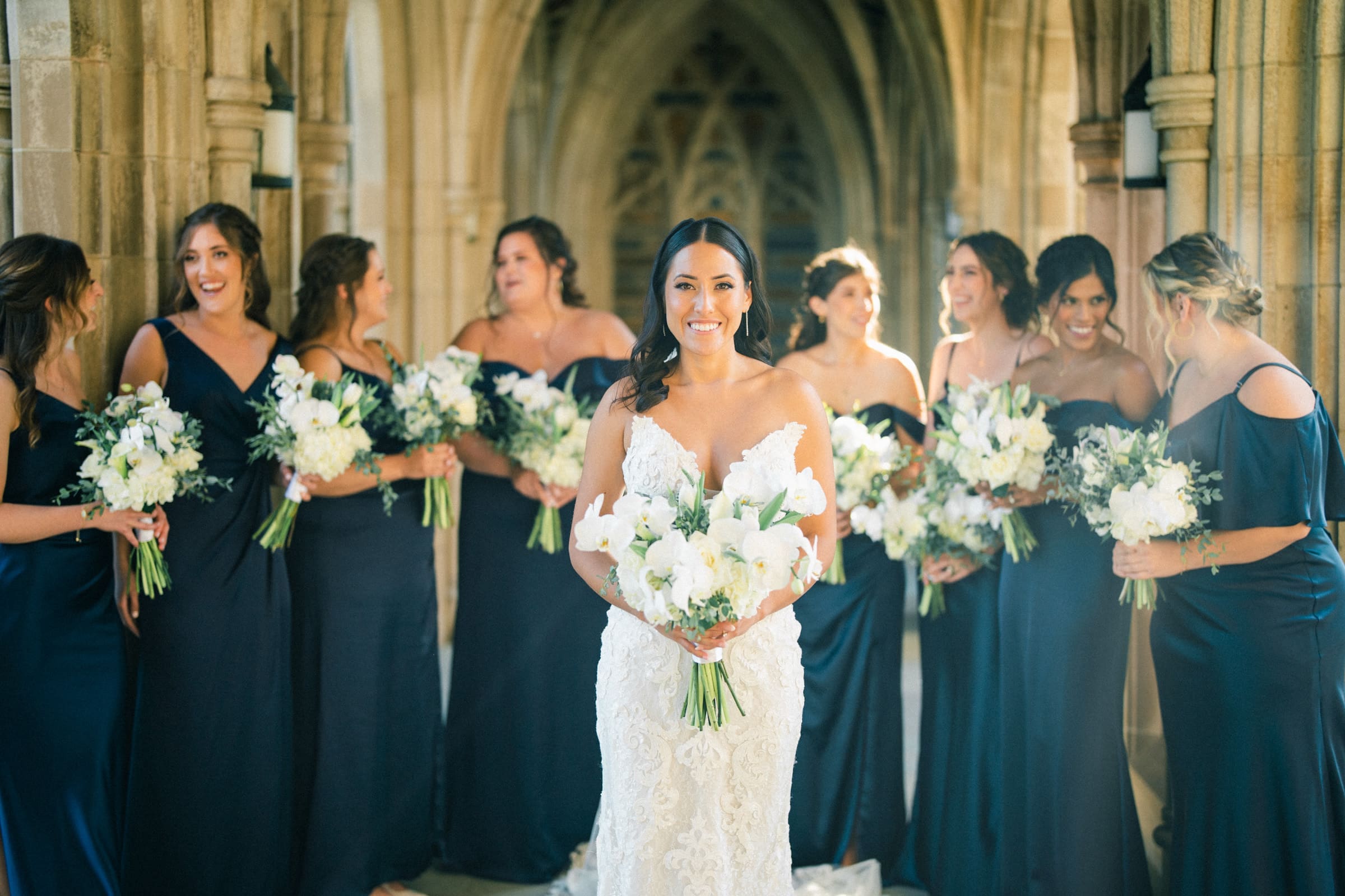 A bride in a white lace gown stands smiling at the center, holding a bouquet of white flowers. She is surrounded by bridesmaids in navy blue dresses, each with similar bouquets, set in the sunlit arches of Duke Gardens—a perfect wedding scene.
