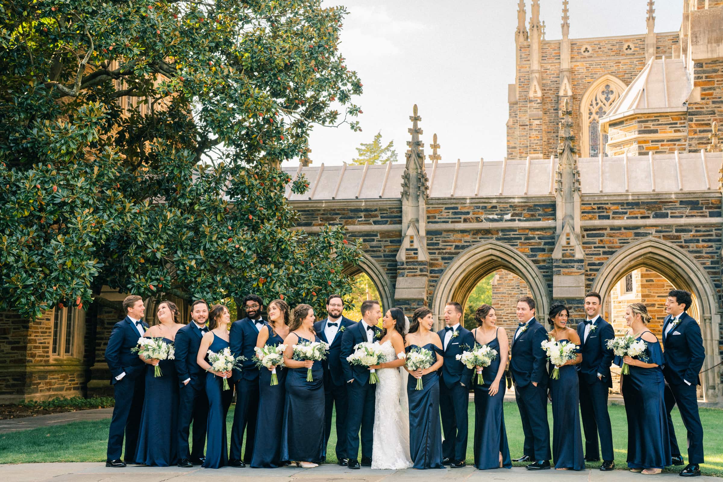 A wedding party poses outdoors in formal attire at the picturesque Duke Gardens. The group, consisting of bridesmaids and groomsmen in dark suits and dresses, holds vibrant bouquets. They stand gracefully before a stone building with arches entwined in lush greenery.