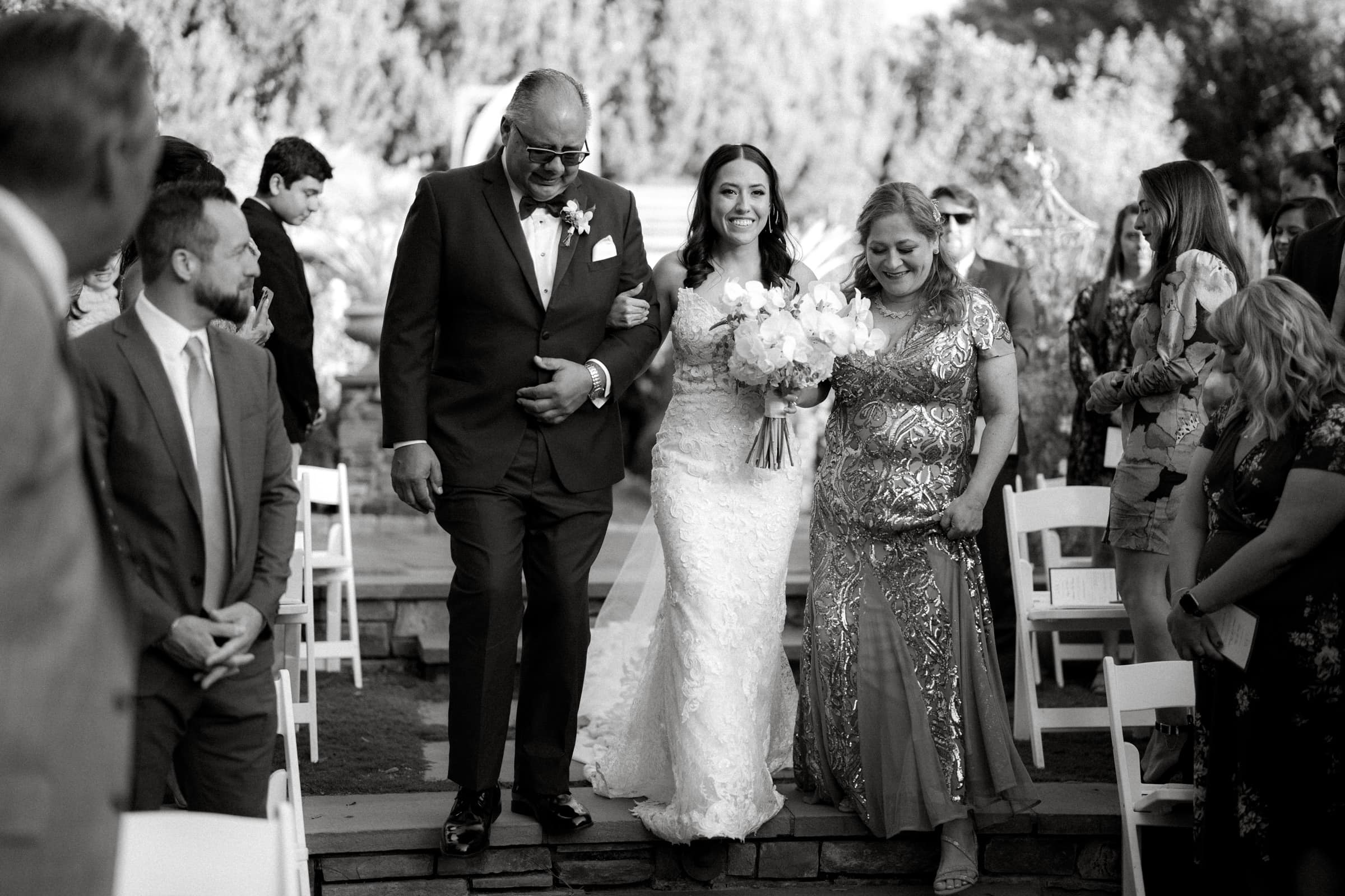 A bride in a lace gown walks down the aisle at Duke Gardens, smiling, arm-in-arm with an older man and woman. They are surrounded by seated guests in an enchanting outdoor setting with trees in the background.