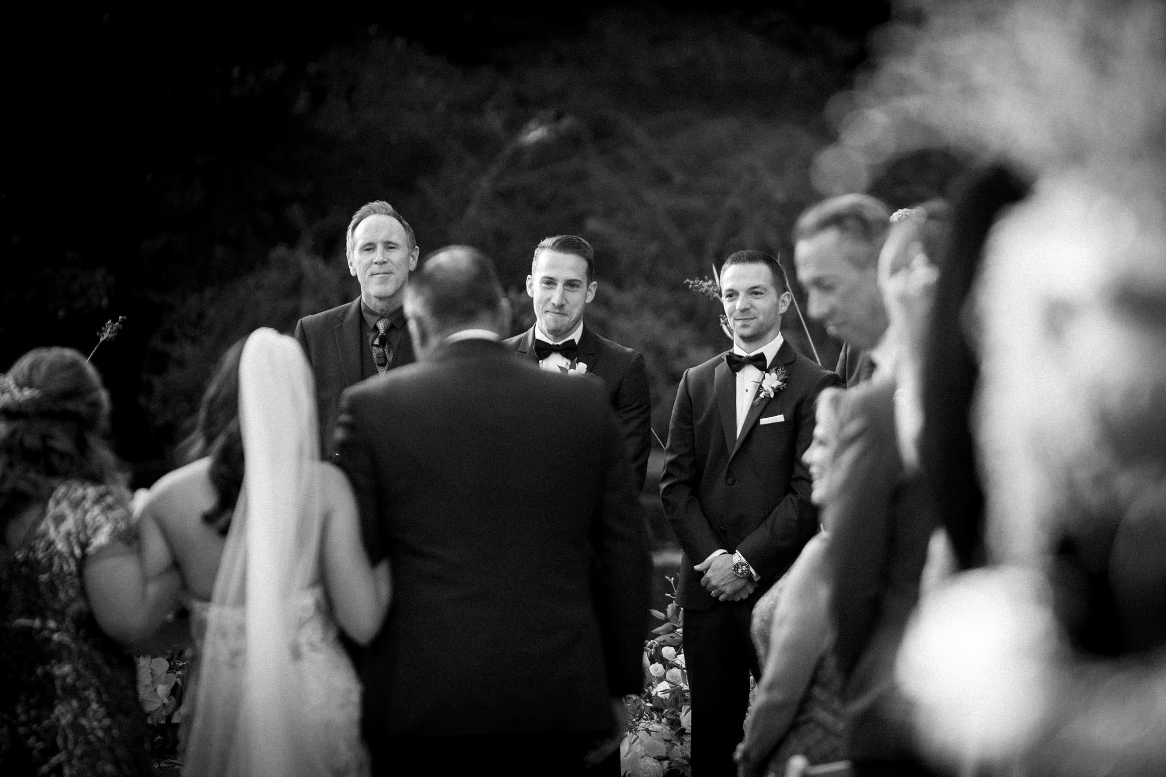 A black-and-white photo captures a Duke Gardens wedding ceremony. The bride, with her back to the camera, walks down the aisle with an escort towards the groom and officiant at the altar. Guests are seated on either side, observing this timeless scene.
