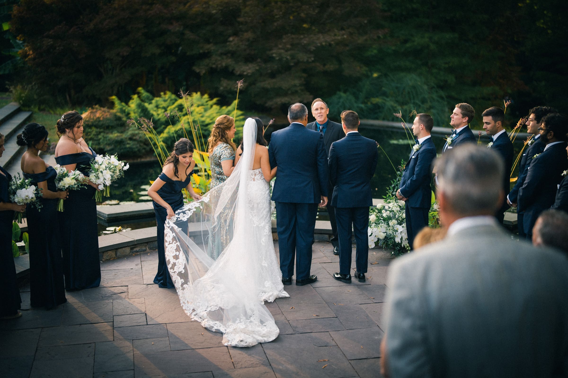 A Duke Gardens wedding unfolds in a picturesque setting. A bride in a white gown with a long veil stands with the groom before an officiant. Bridesmaids and groomsmen in formal wear surround them, as guests watch from nearby, embraced by the lush greenery of the gardens.