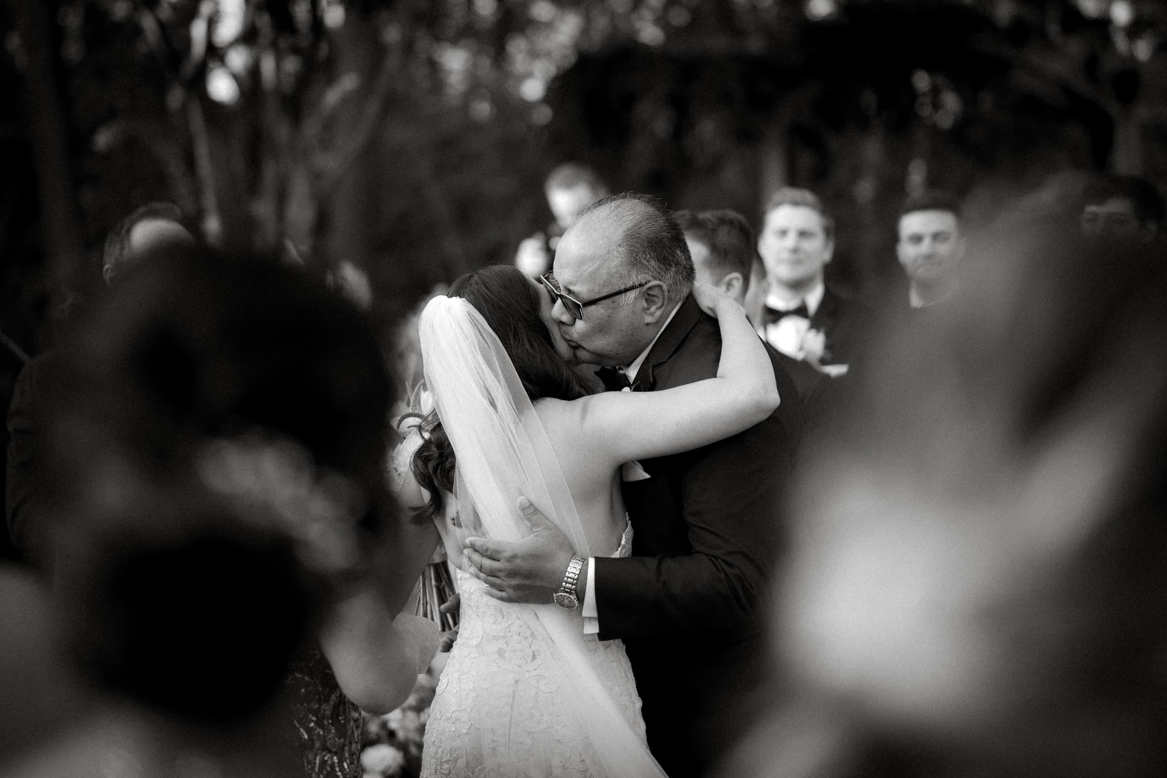A bride in a white wedding dress and veil embraces an older man in a suit and glasses during an outdoor ceremony at Duke Gardens. The background, with its blurred guests and trees, captures a warm, emotional moment.