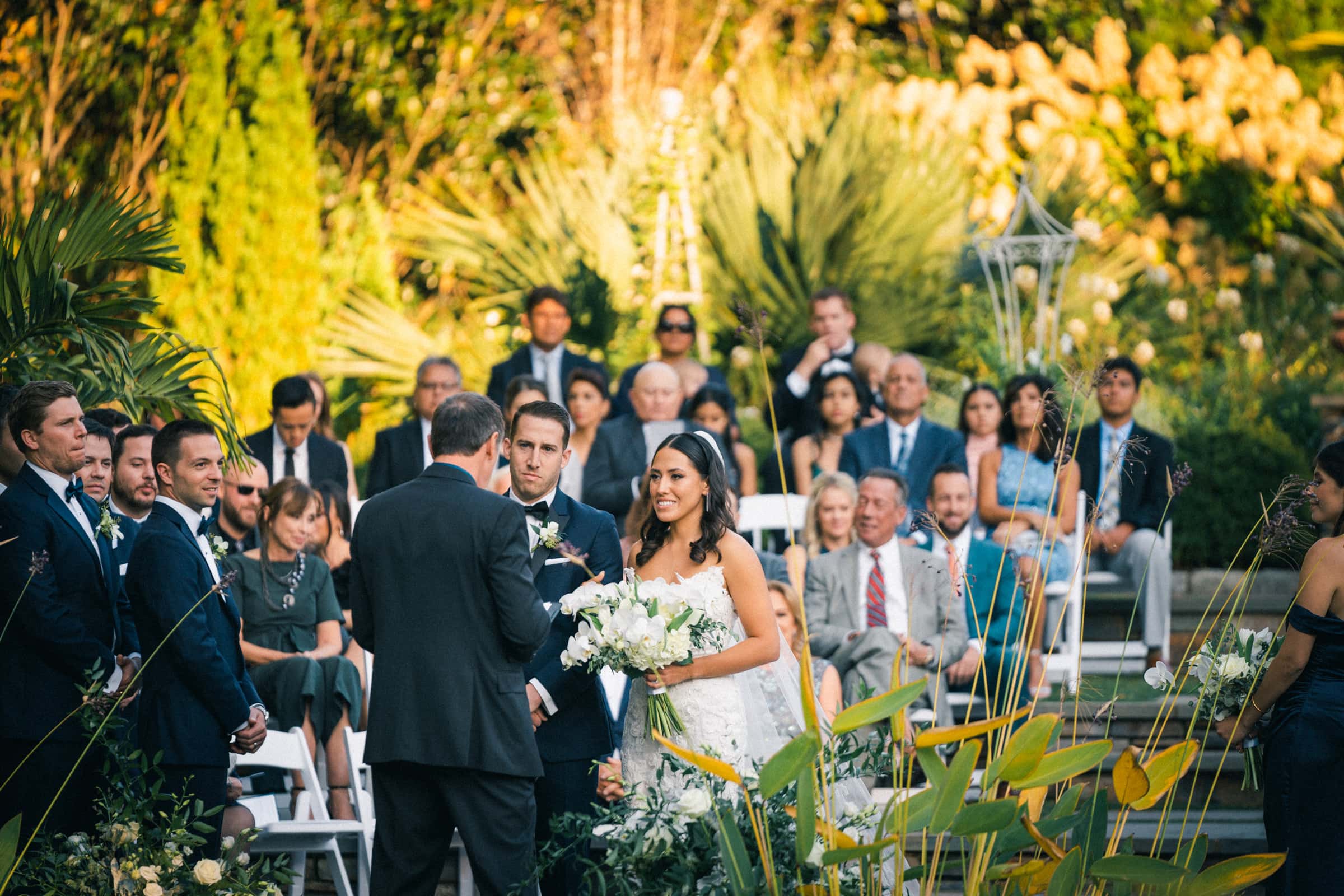 A bride and groom stand together at a beautiful Duke Gardens wedding ceremony. The bride holds a bouquet of white flowers, while guests are seated in the background, surrounded by lush greenery and plants under the warm sunlight.
