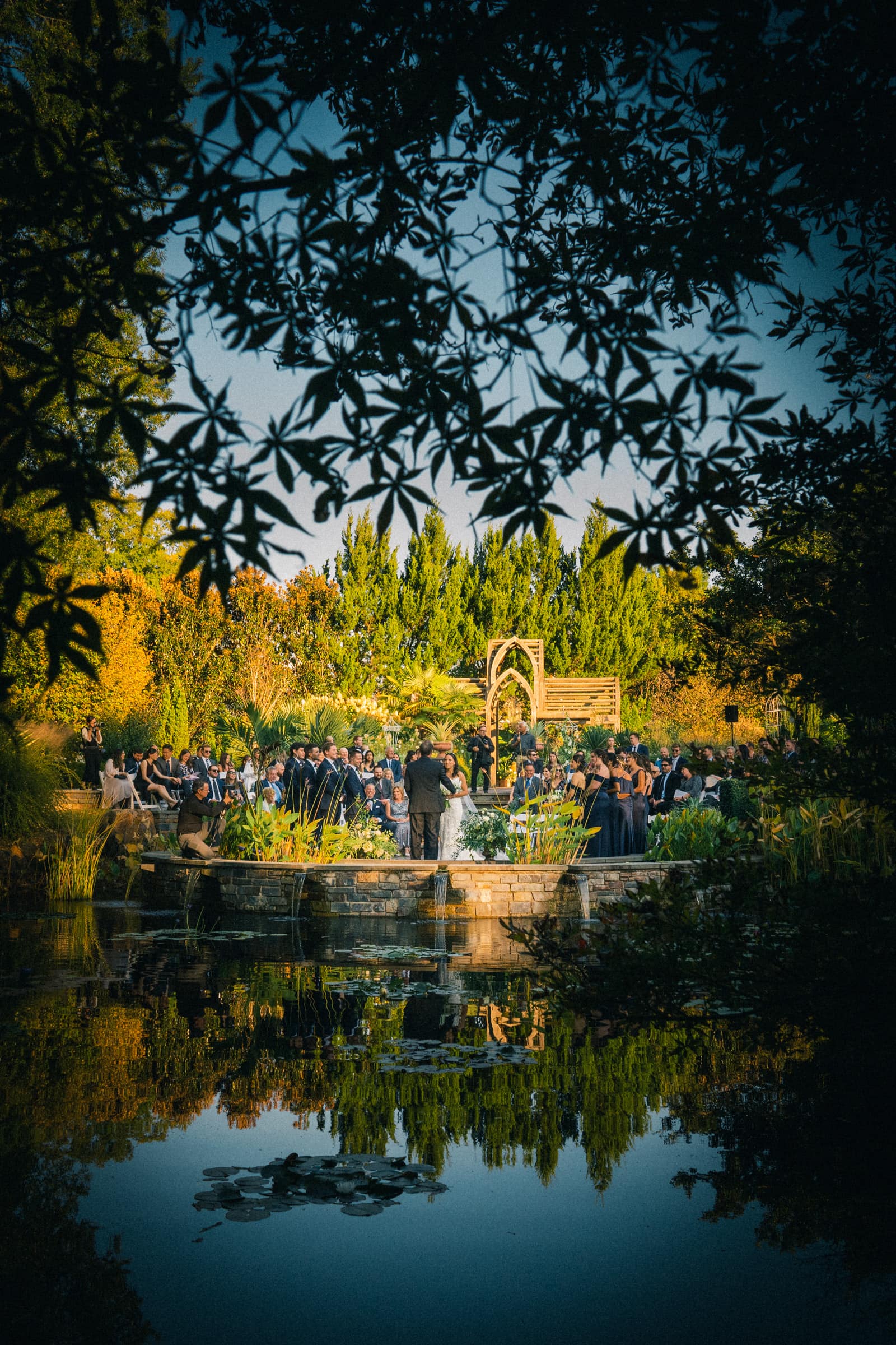 A serene outdoor wedding ceremony at Duke Gardens, viewed through foliage. Guests seated around a stone path surrounded by greenery and a pond reflecting the scene. A wooden arch serves as the altar under the clear blue sky.