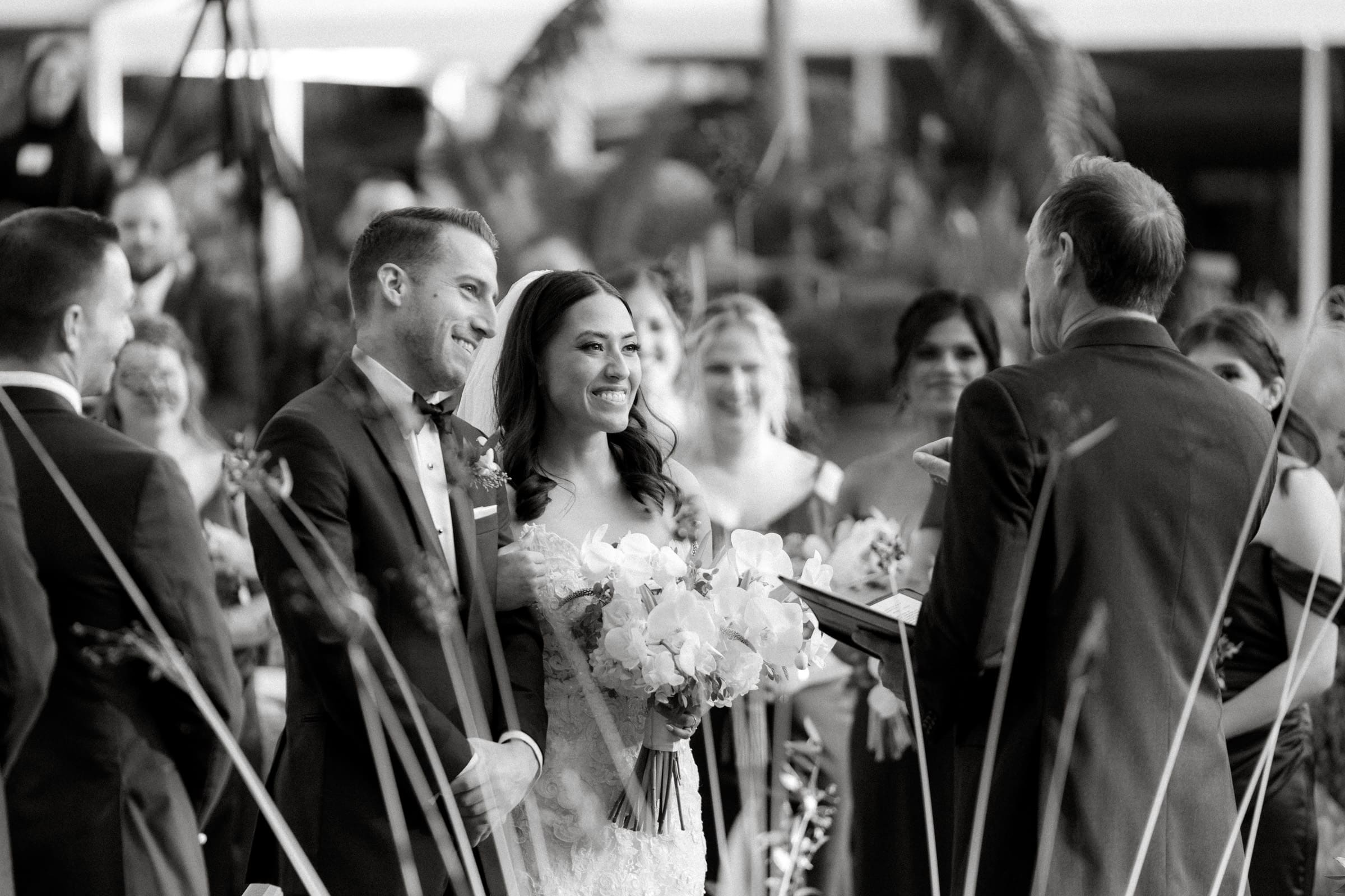A black and white photo captures a Duke Gardens wedding ceremony. The bride and groom stand smiling, holding hands. The groom wears a suit while the bride holds a bouquet. The officiant speaks, surrounded by bridesmaids and groomsmen in the lush garden backdrop.