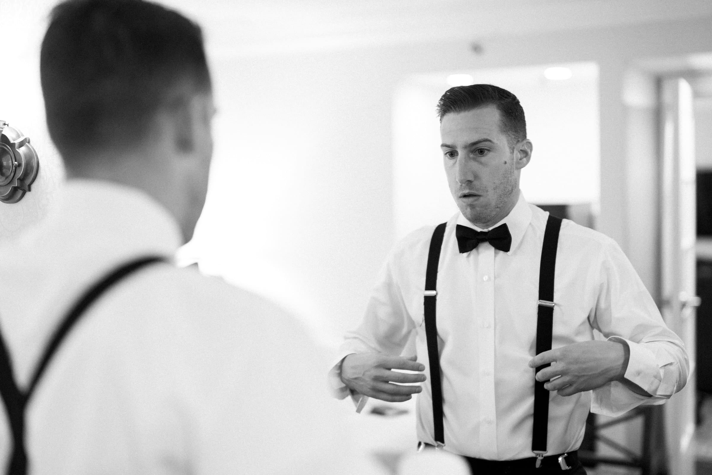 In a black and white photo, a man in a white shirt, black suspenders, and bow tie stands before a mirror, adjusting his attire with focused intent. The well-lit room hints at the elegance of Duke Gardens wedding preparation. He is captured from the waist up.