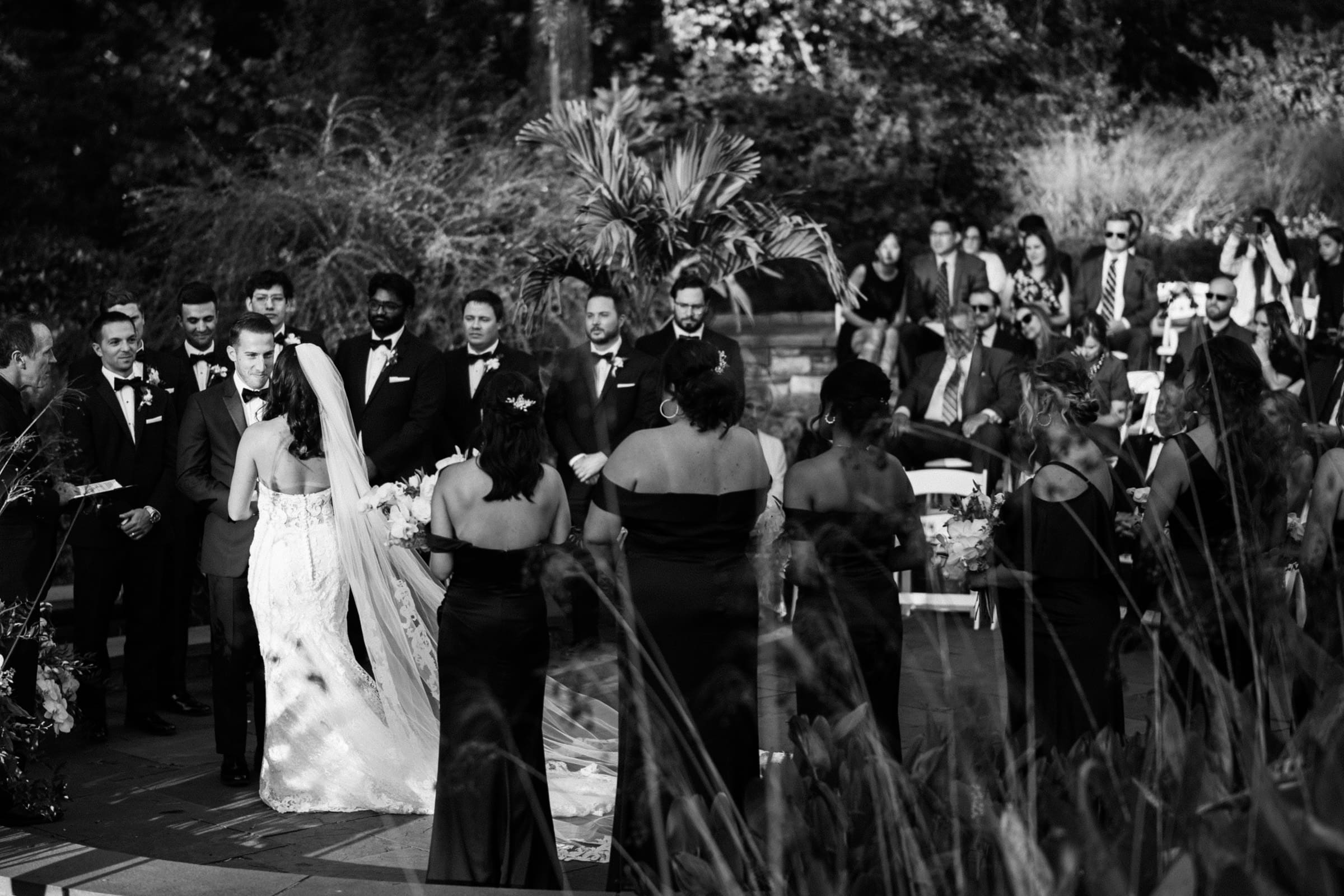 Black and white photo of an elegant outdoor wedding ceremony at Duke Gardens. The bride and groom stand facing each other, surrounded by bridesmaids and groomsmen. Guests are seated nearby, with trees and leaves providing a picturesque backdrop.