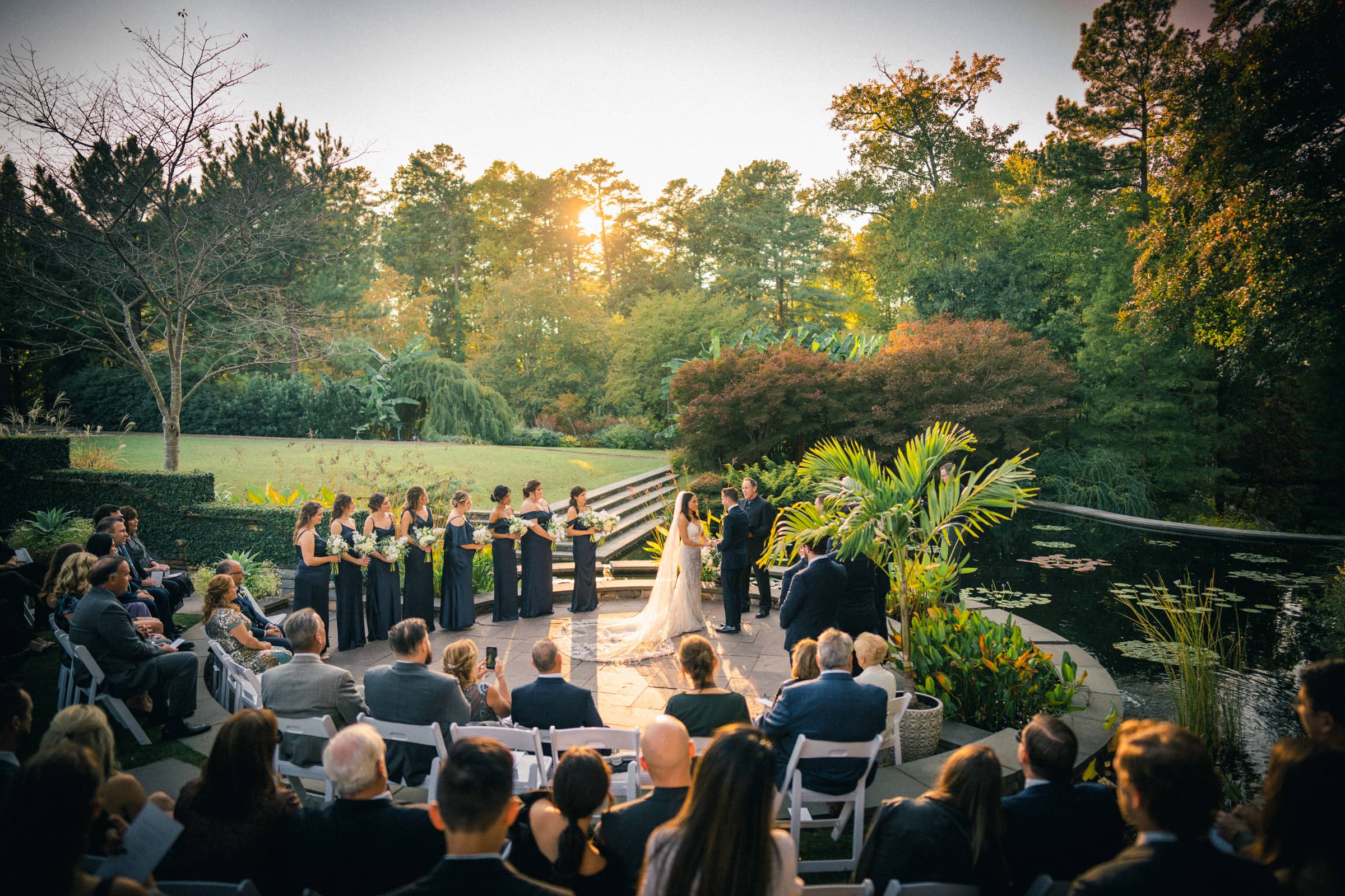 A Duke Gardens wedding at sunset unfolds in a lush garden setting. The couple stands before the officiant, bridal party on either side, as guests sit surrounded by greenery and a serene pond with lily pads. Sunlight filters through the trees, enhancing the magical atmosphere.