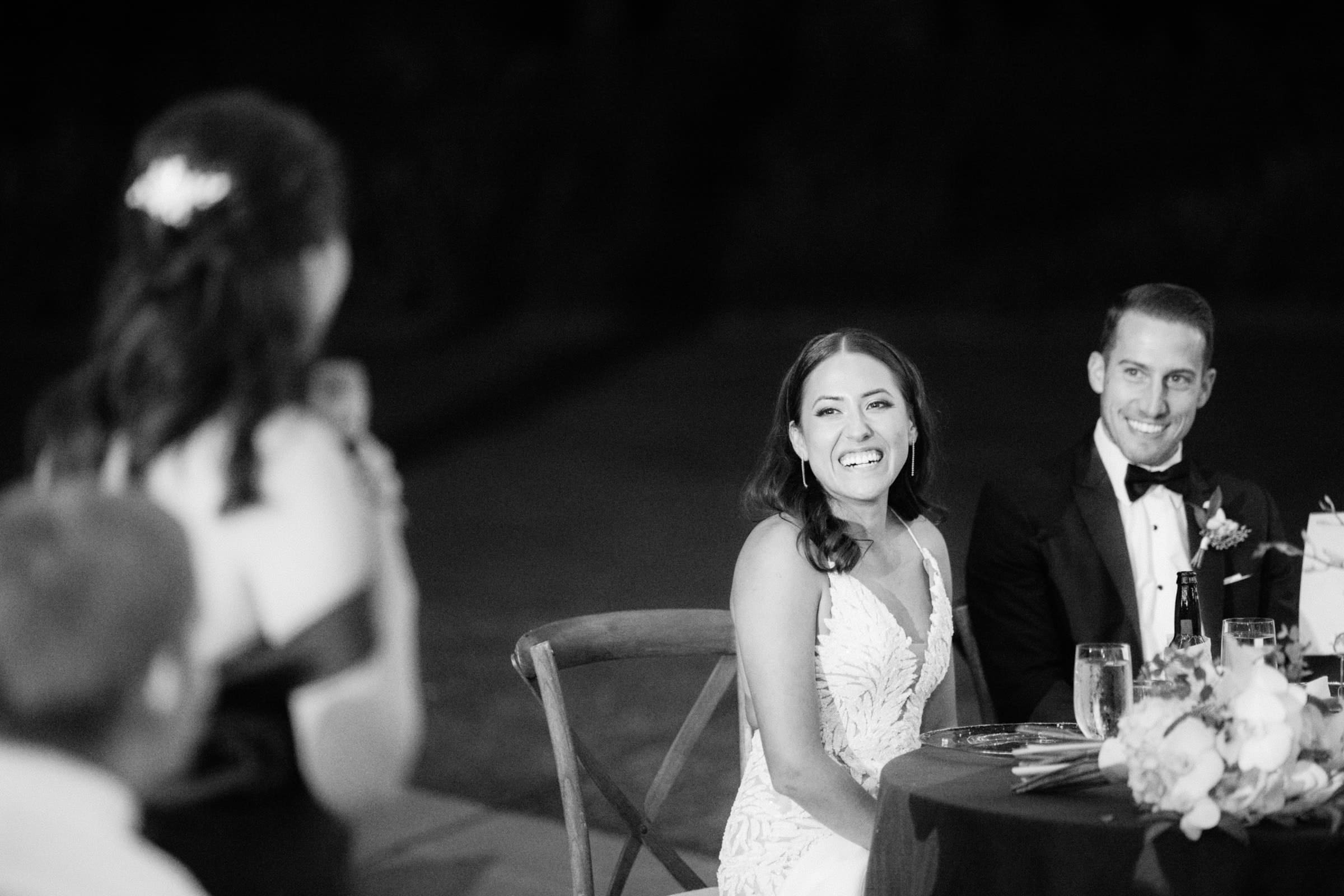 At a Duke Gardens wedding, the bride and groom sit at a table, smiling as they listen to a heartfelt speech. The bride dazzles in her white gown, while the groom looks dapper in his tuxedo. In the foreground, a woman raises her glass for a toast amidst the warm, festive atmosphere.