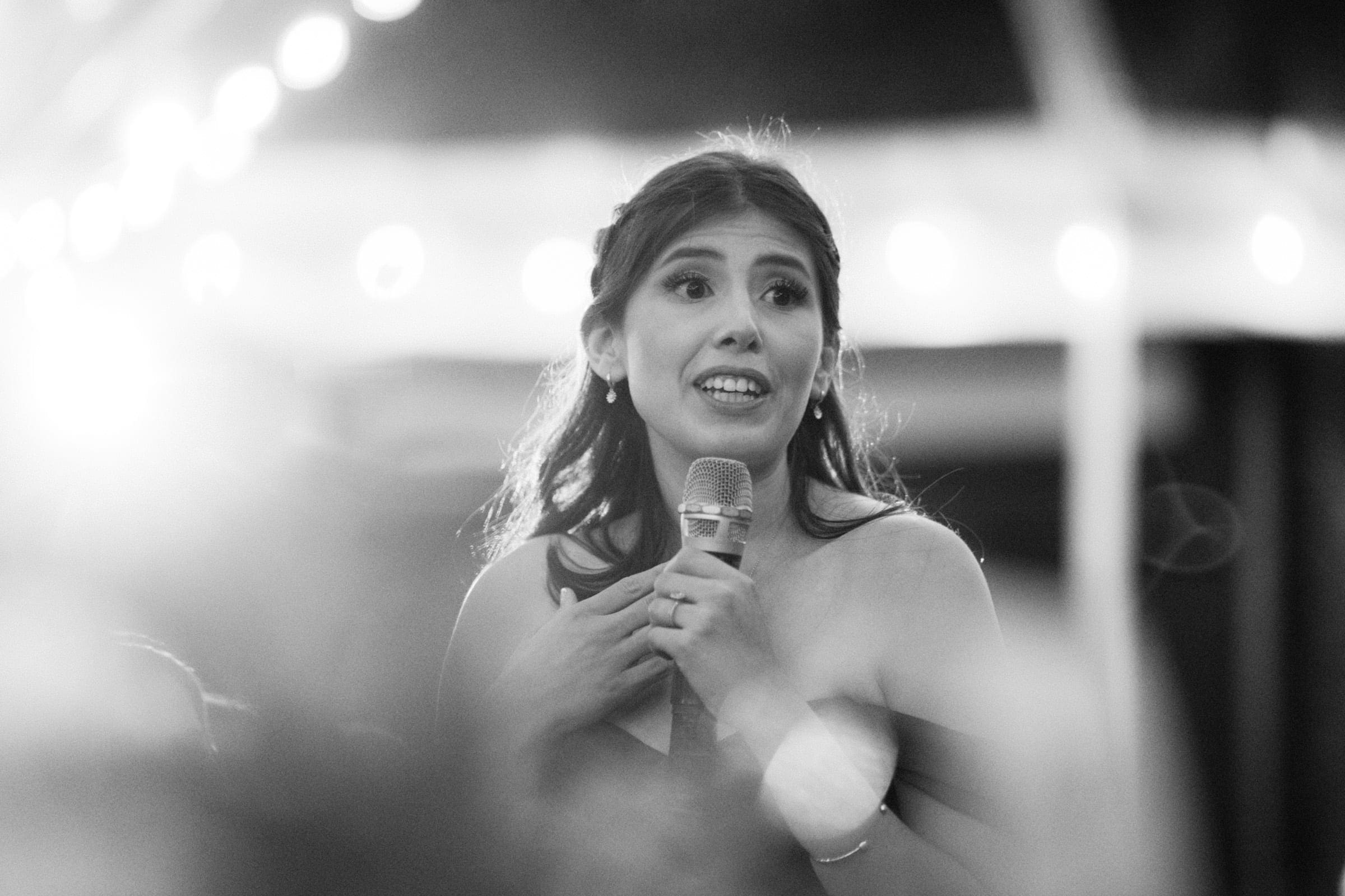 A woman in a dark dress speaks into a microphone with a heartfelt expression, as if at a Duke Gardens wedding. The black and white photo captures the glow of lights in the background, creating a warm and intimate atmosphere.