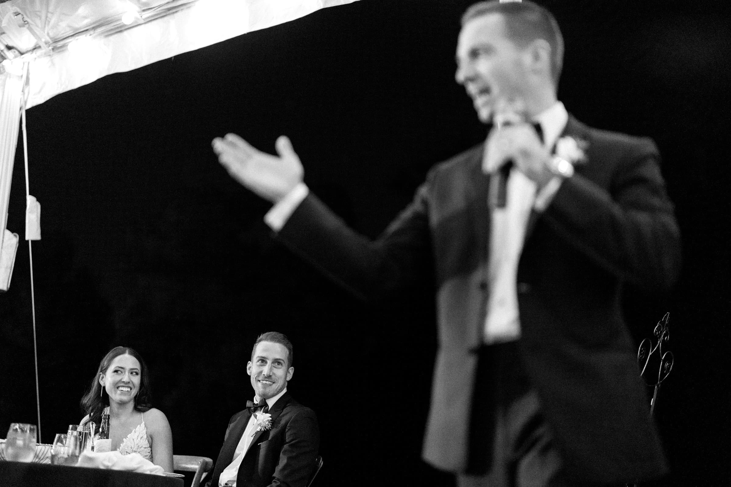 A man in a suit is speaking into a microphone, gesturing animatedly at what seems to be a Duke Gardens wedding. In the background, a seated couple, dressed formally, are smiling and watching him at this elegant event.