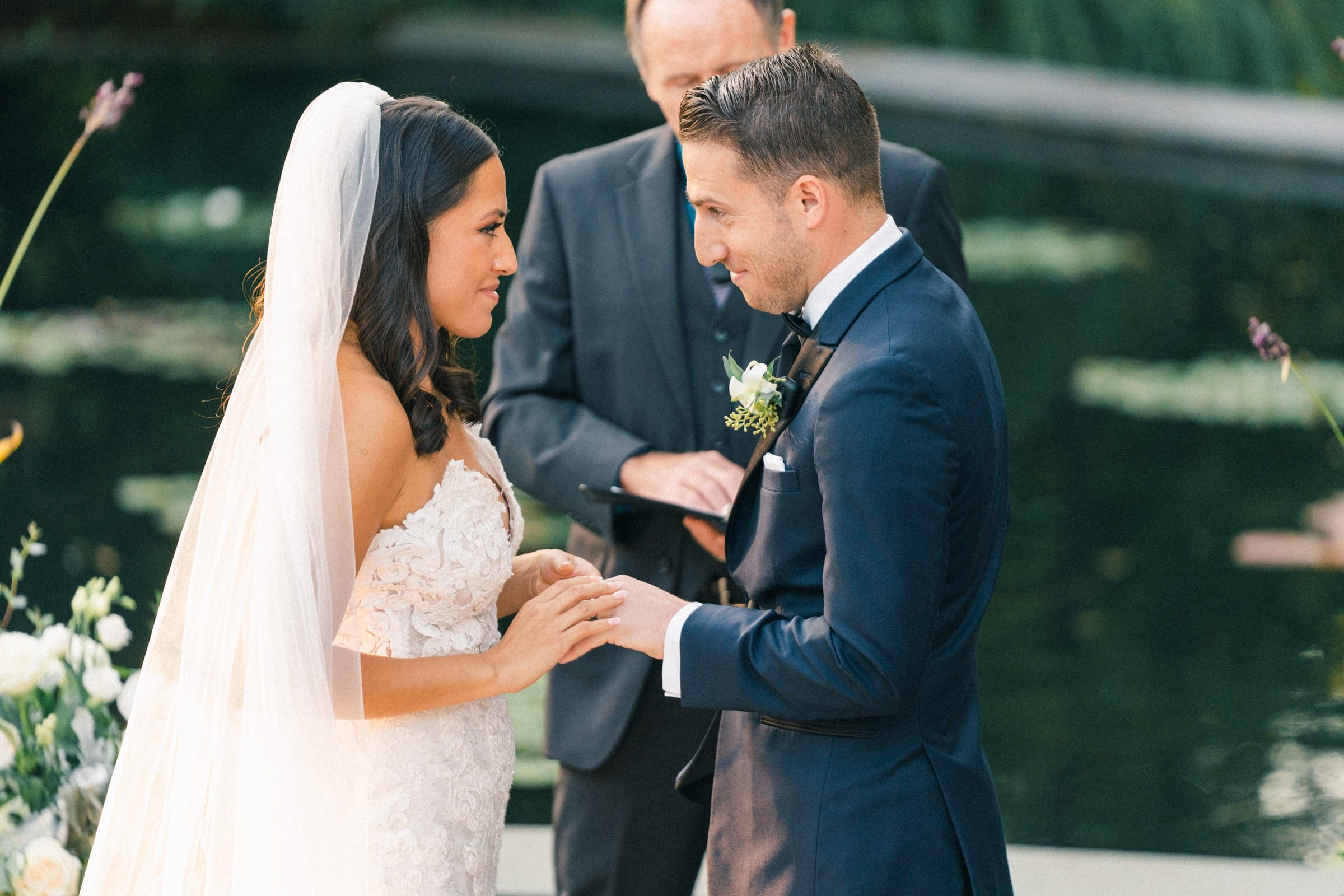 At a Duke Gardens wedding, a bride and groom stand facing each other, holding hands during their outdoor ceremony. The bride wears a white gown and veil, while the groom is in a dark suit. Serene water and lush greenery provide the perfect backdrop for their vows.