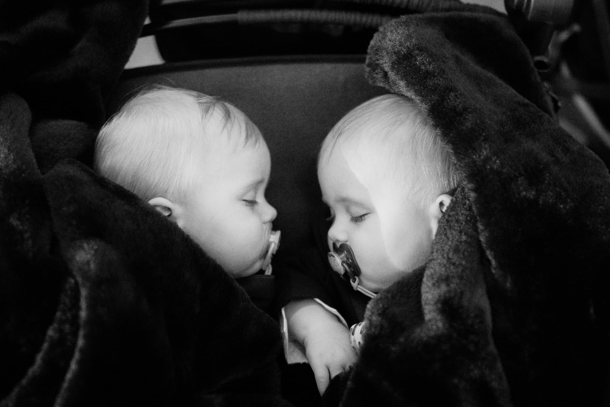 Two babies peacefully sleeping side by side under a dark, plush blanket evoke the serene elegance of a Duke Gardens wedding. Both have pacifiers in their mouths. The image is in black and white, adding a timeless feel to this calming scene.