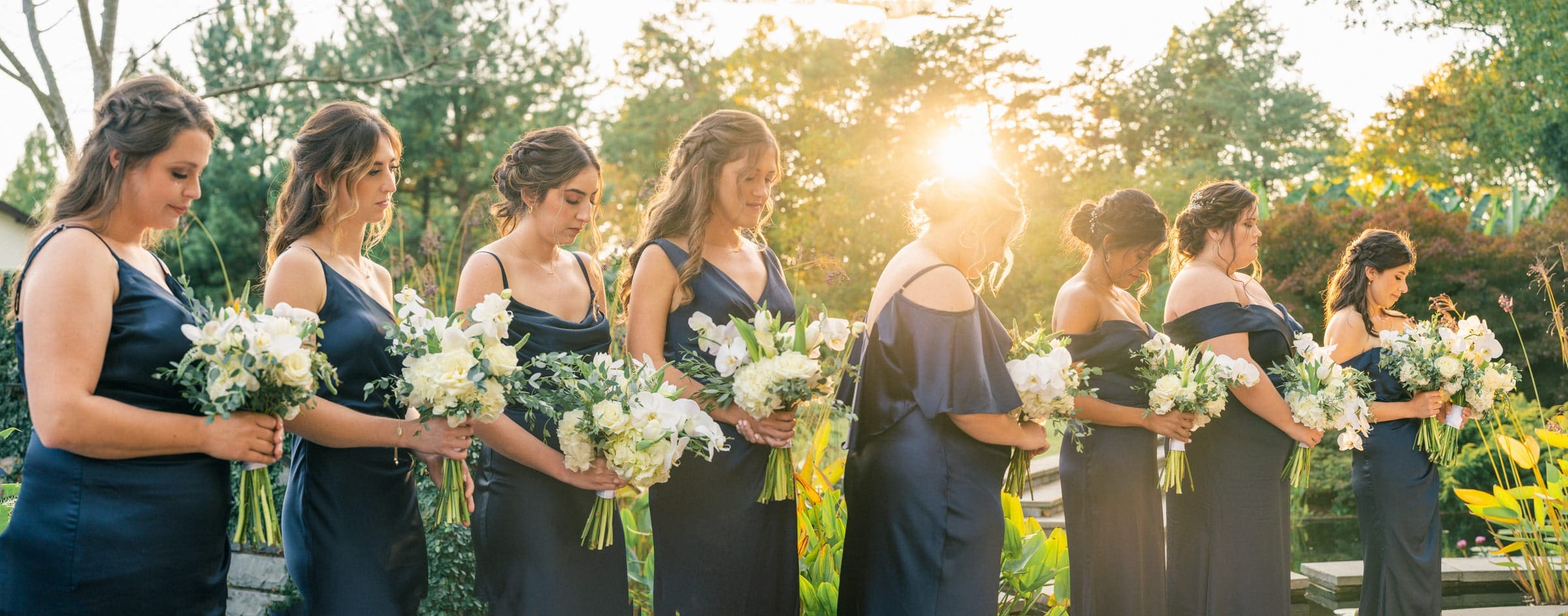 A group of bridesmaids in matching dark blue dresses stands in Duke Gardens, holding white floral bouquets. They are lined up in a row, with the sun setting behind them, creating a warm, glowing atmosphere at this picturesque wedding venue.