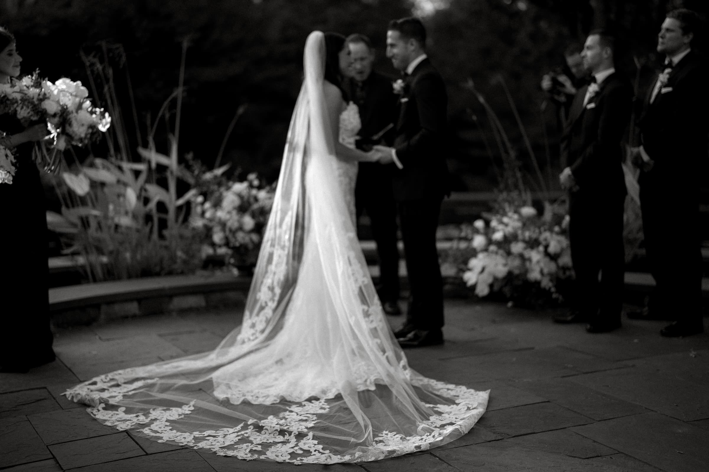 A bride and groom stand facing each other during a Duke Gardens wedding. The bride wears a long, flowing lace veil, while the groom is in a dark suit. They are surrounded by groomsmen and floral decorations on the stone patio.