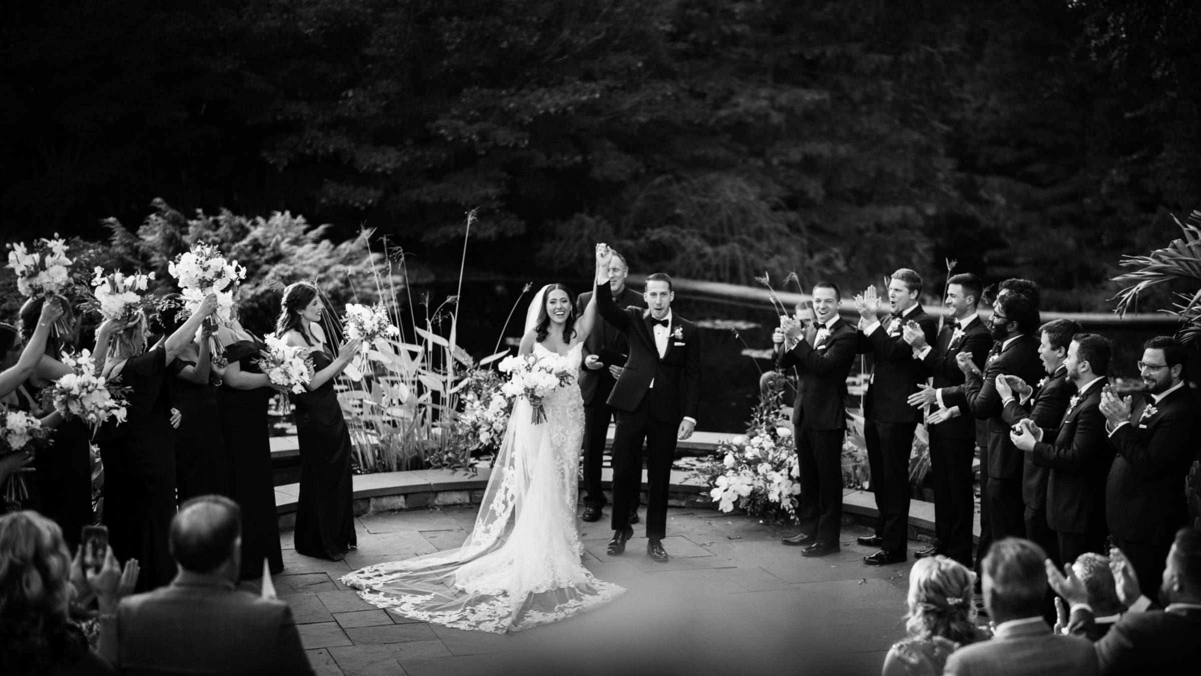 A black and white photo captures a bride and groom holding hands at their Duke Gardens wedding. The bride wears a long, lace gown. Bridesmaids and groomsmen stand around them, clapping, as guests seated in the foreground share in this enchanting moment.